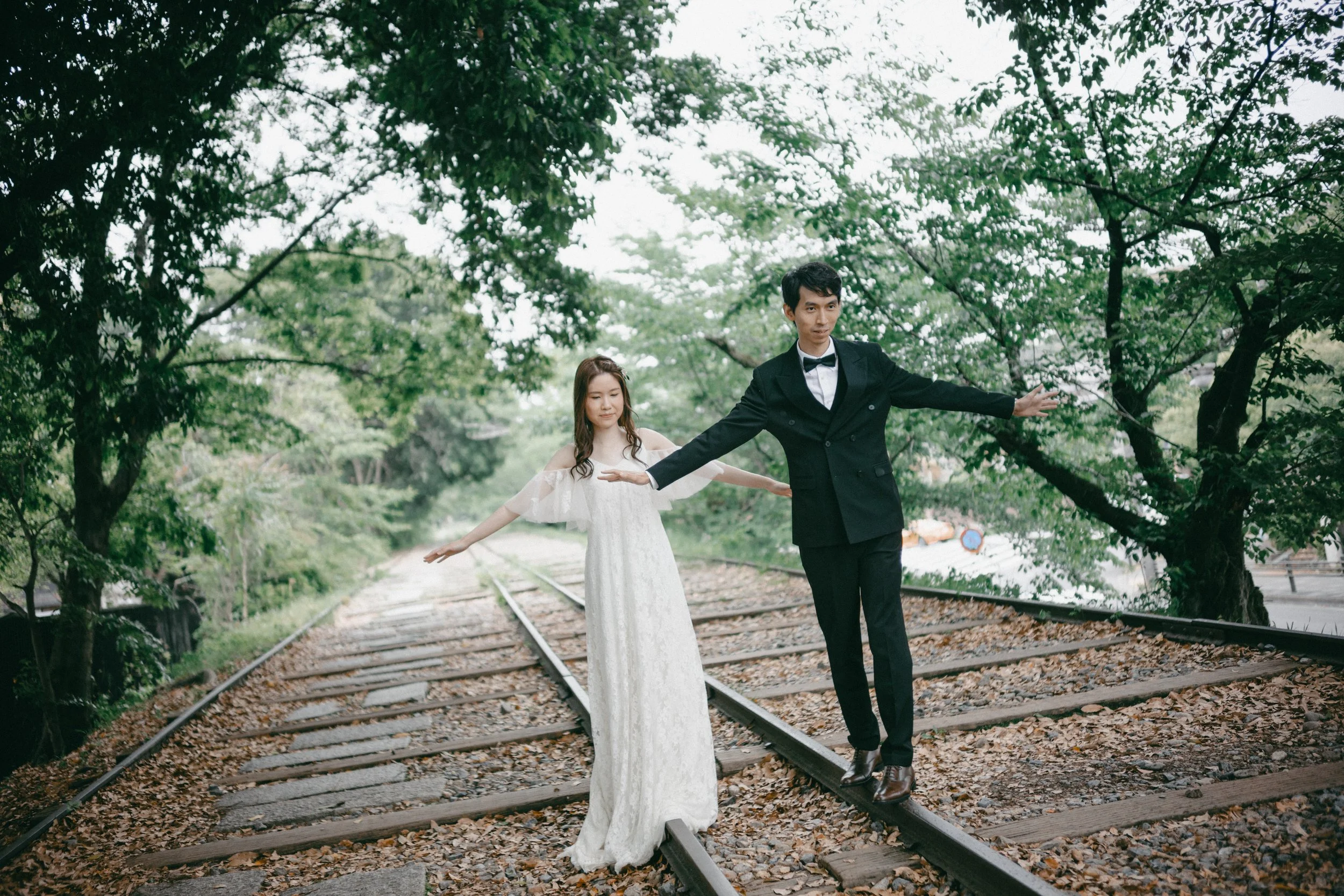 A man and woman in wedding attire balancing on train tracks surrounded by green trees, with arms extended.