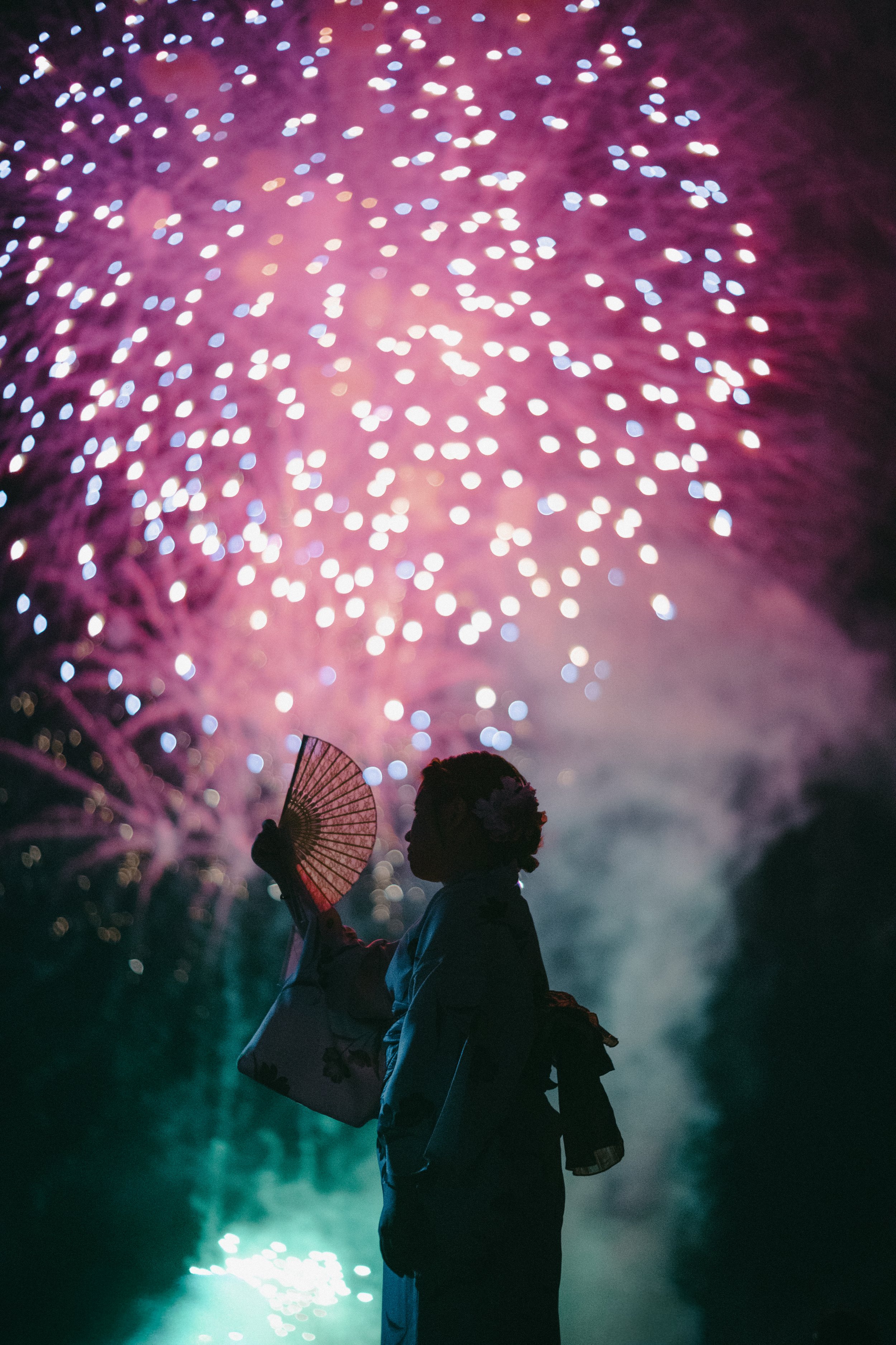 A silhouette of a woman wearing a traditional kimono and holding a paper fan, watching fireworks explode in the night sky with pink, blue, and purple colors.