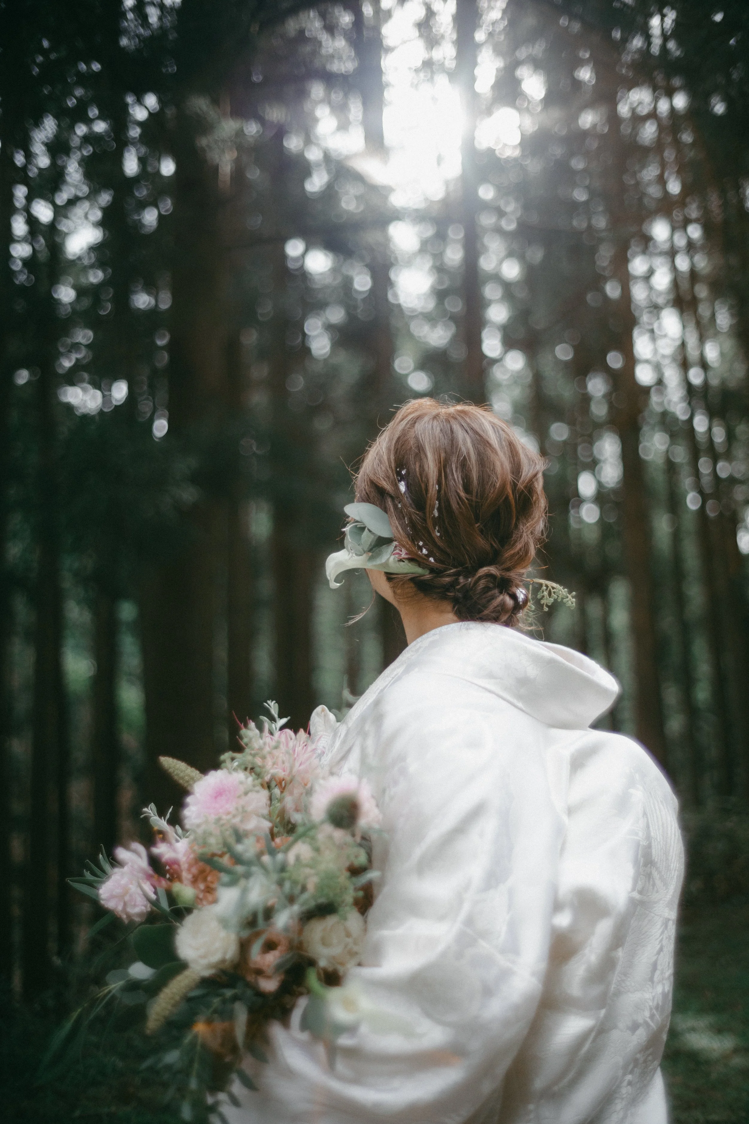 A woman with brown hair styled in an updo, holding a bouquet of flowers, stands in a forest with sunlight filtering through tall trees.