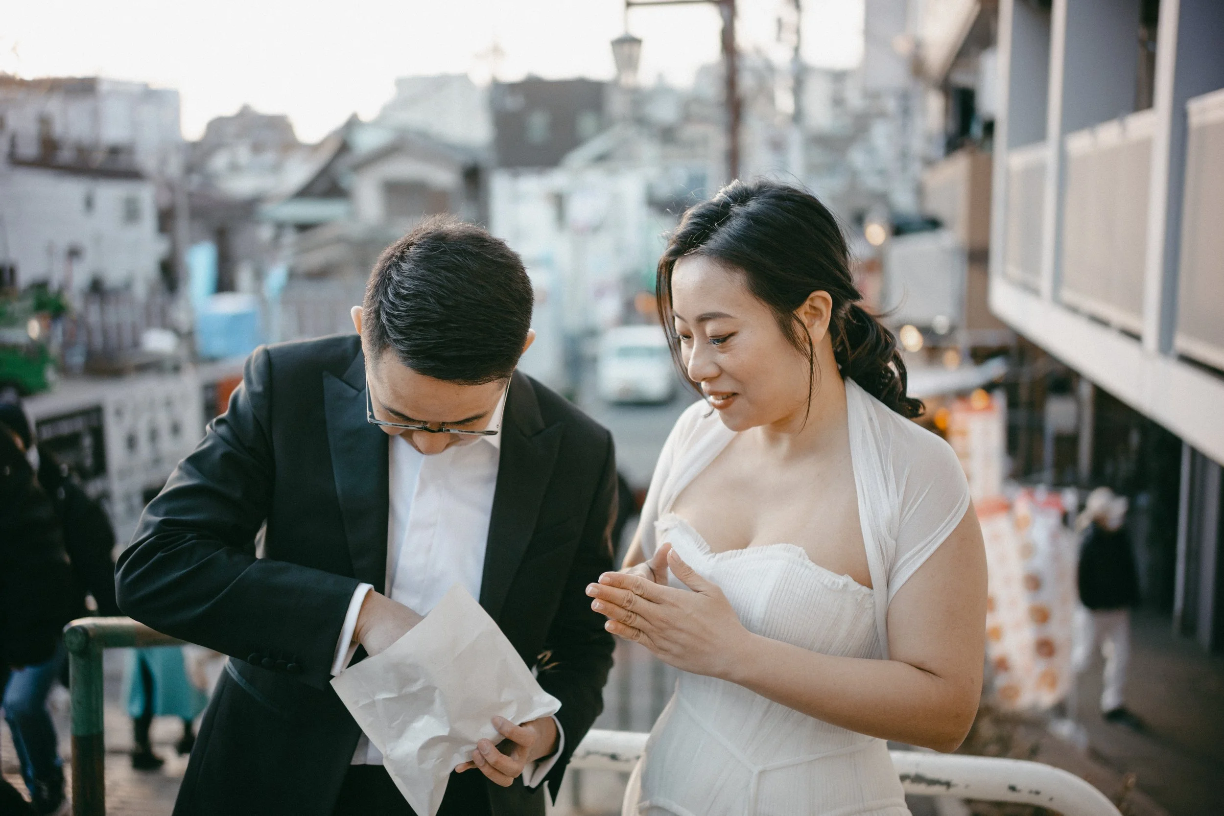 A man in a black suit and glasses and a woman in a white dress are examining a paper bag on a city street during daytime.