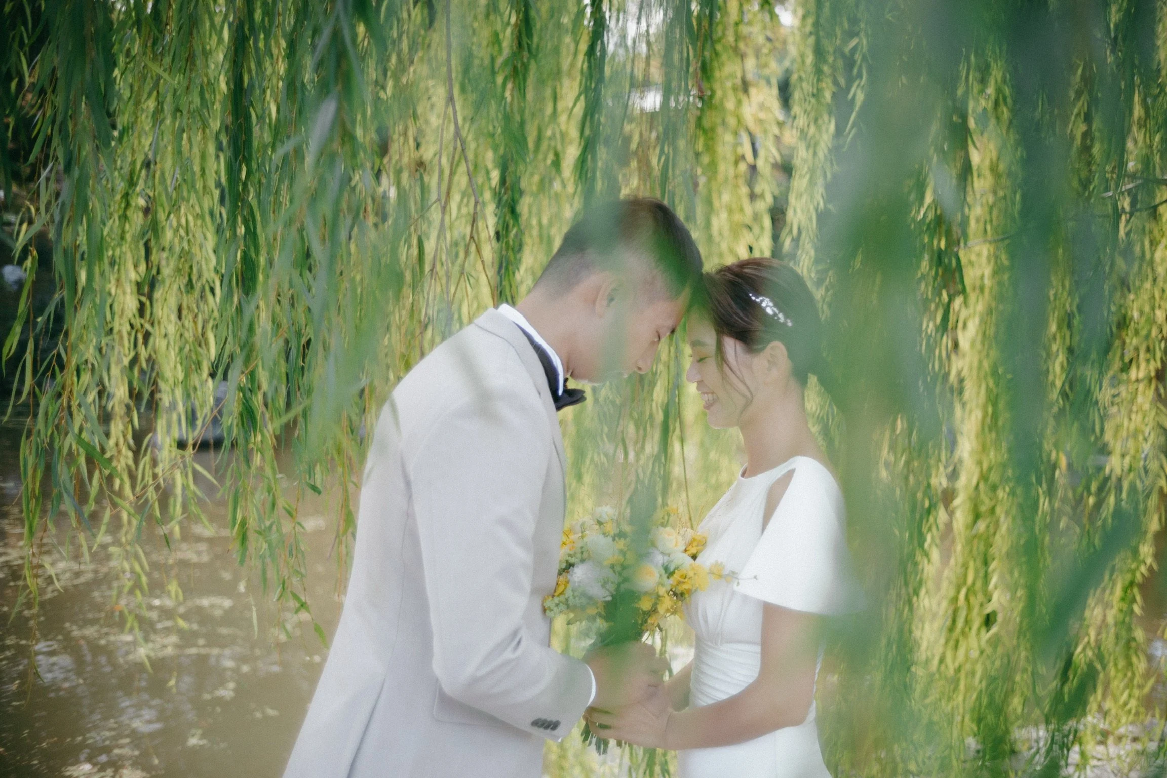 A bride and groom standing close together under hanging green willow branches, with the groom holding a bouquet of yellow flowers and both smiling at each other.