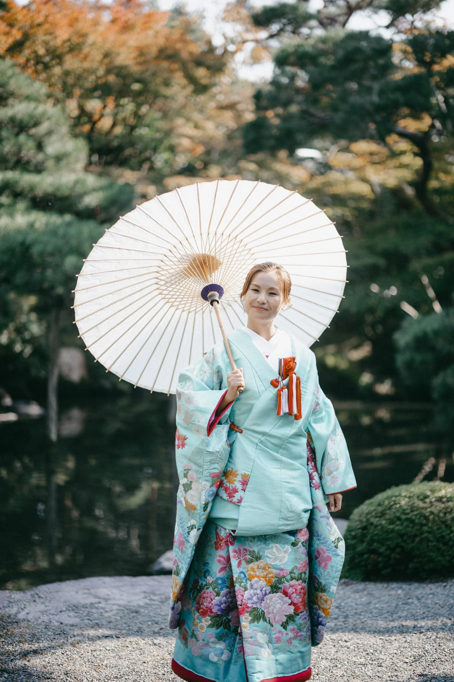 A woman dressed in a colorful traditional Japanese kimono, holding a paper parasol, standing outdoors near a pond with trees in the background.