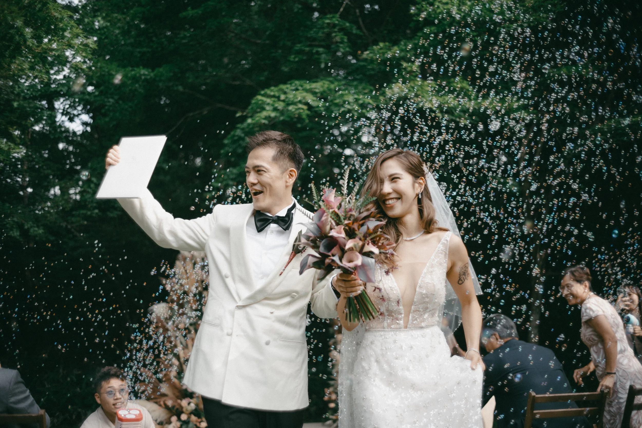 A newly married couple celebrating outdoors with guests, with bubbles floating around, as the groom holds a white certificate or card and the bride holds a bouquet of flowers.