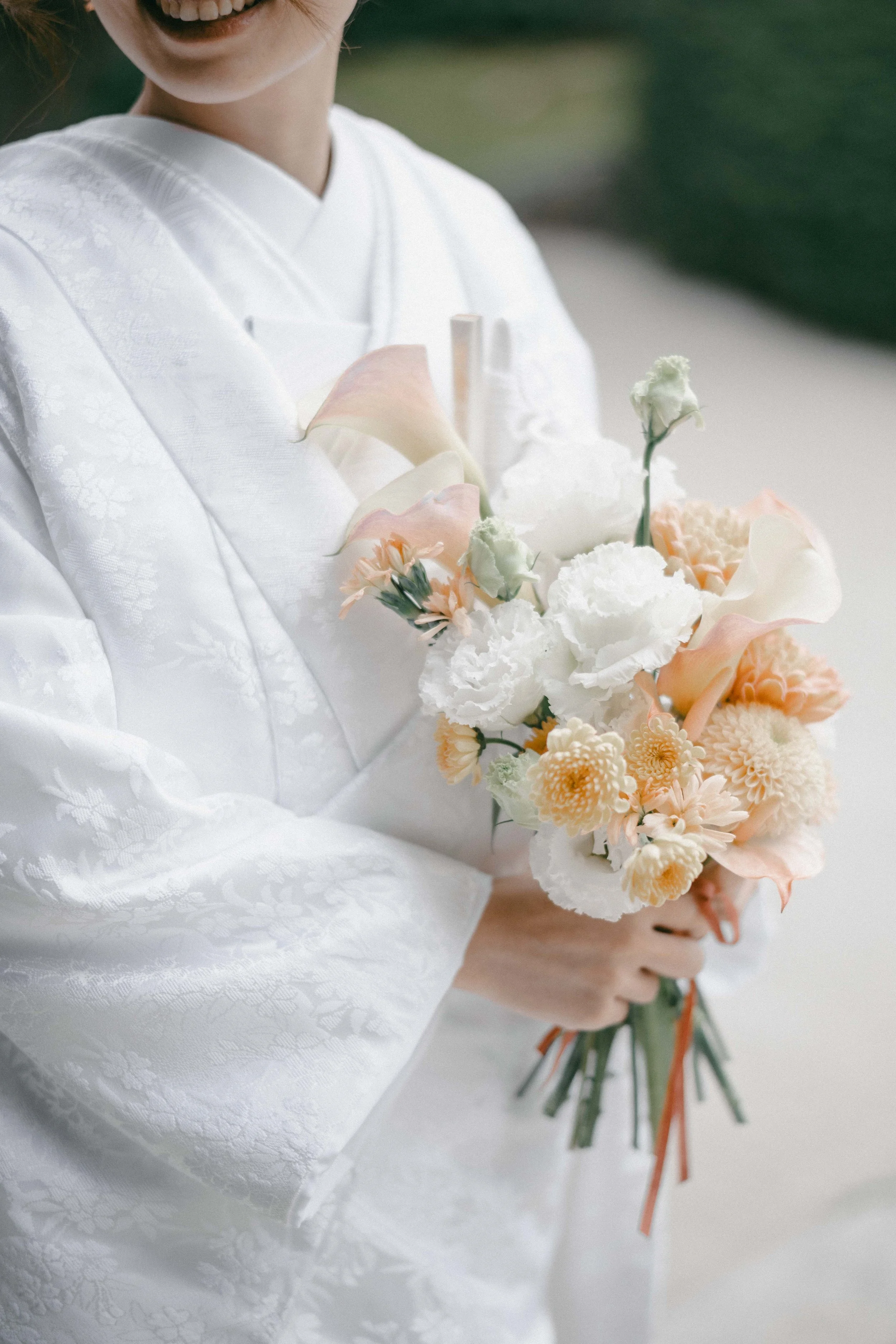 A person in a white, patterned kimono is holding a bouquet of pastel-colored flowers including white, peach, and light pink flowers.