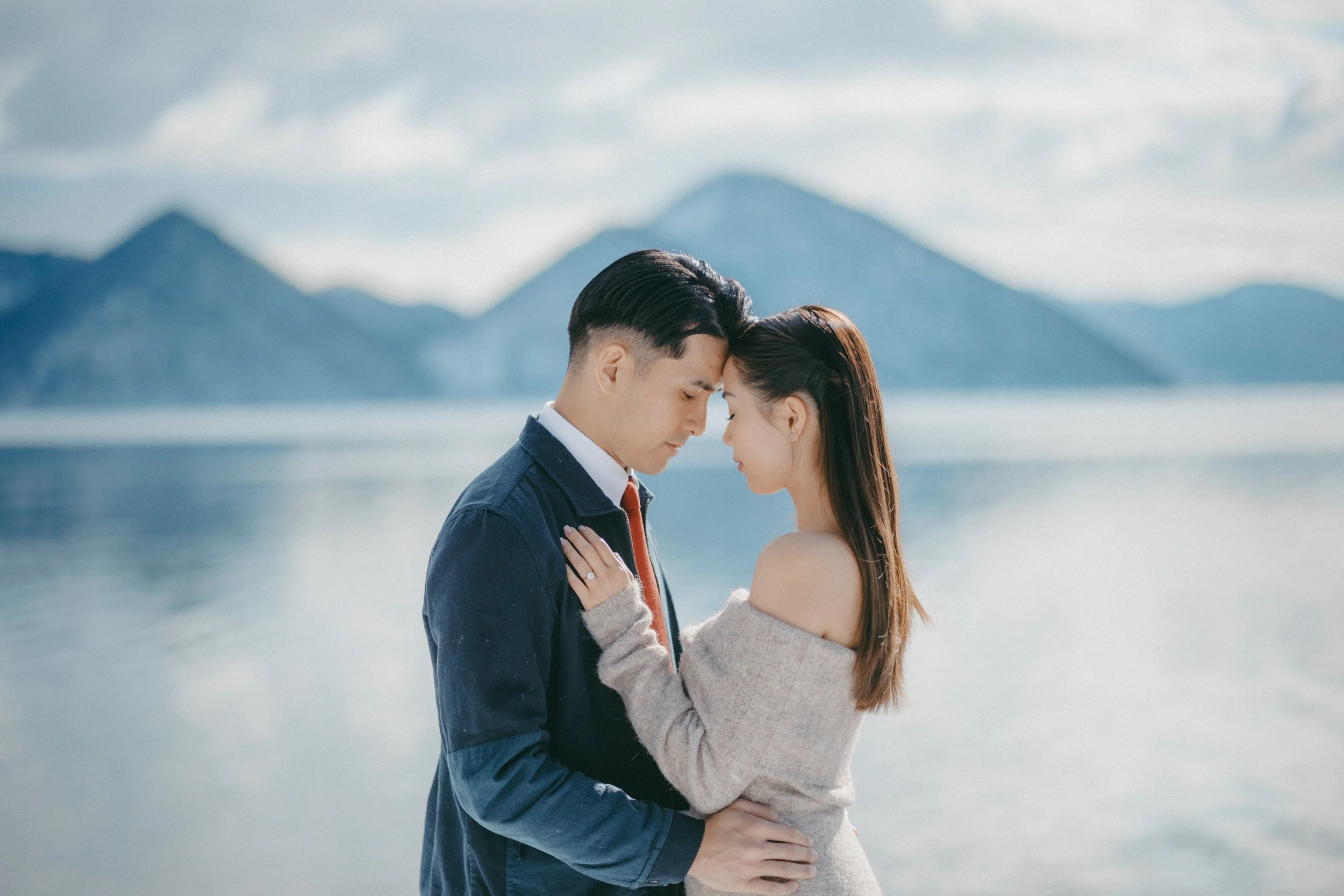 A man and woman sharing an intimate moment by a lake with mountains in the background, touching foreheads with eyes closed.