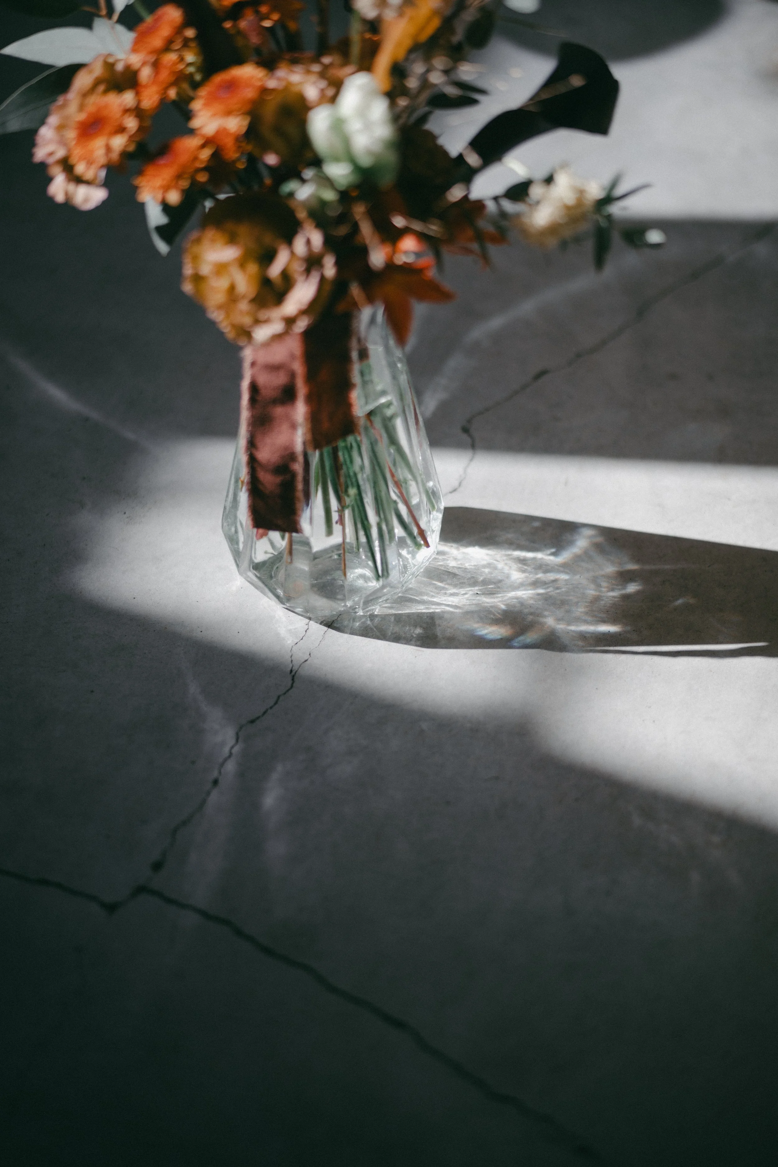 A glass vase with a bouquet of orange, white, and yellow flowers resting on a cracked concrete floor, with sunlight casting a shadow and reflections on the surface.