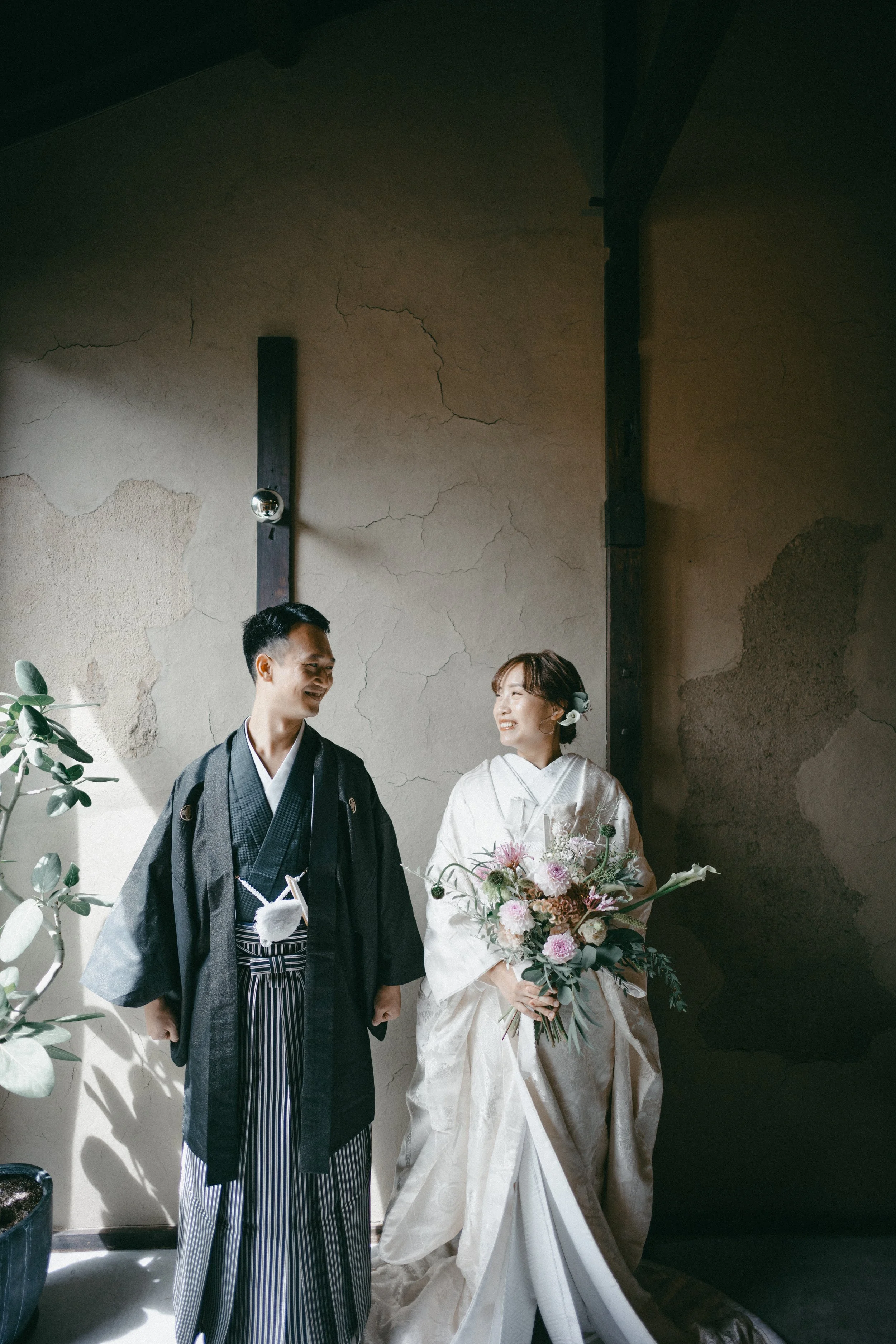 A bride and groom in traditional Japanese wedding attire smiling at each other indoors with a textured wall and potted plant in the background.