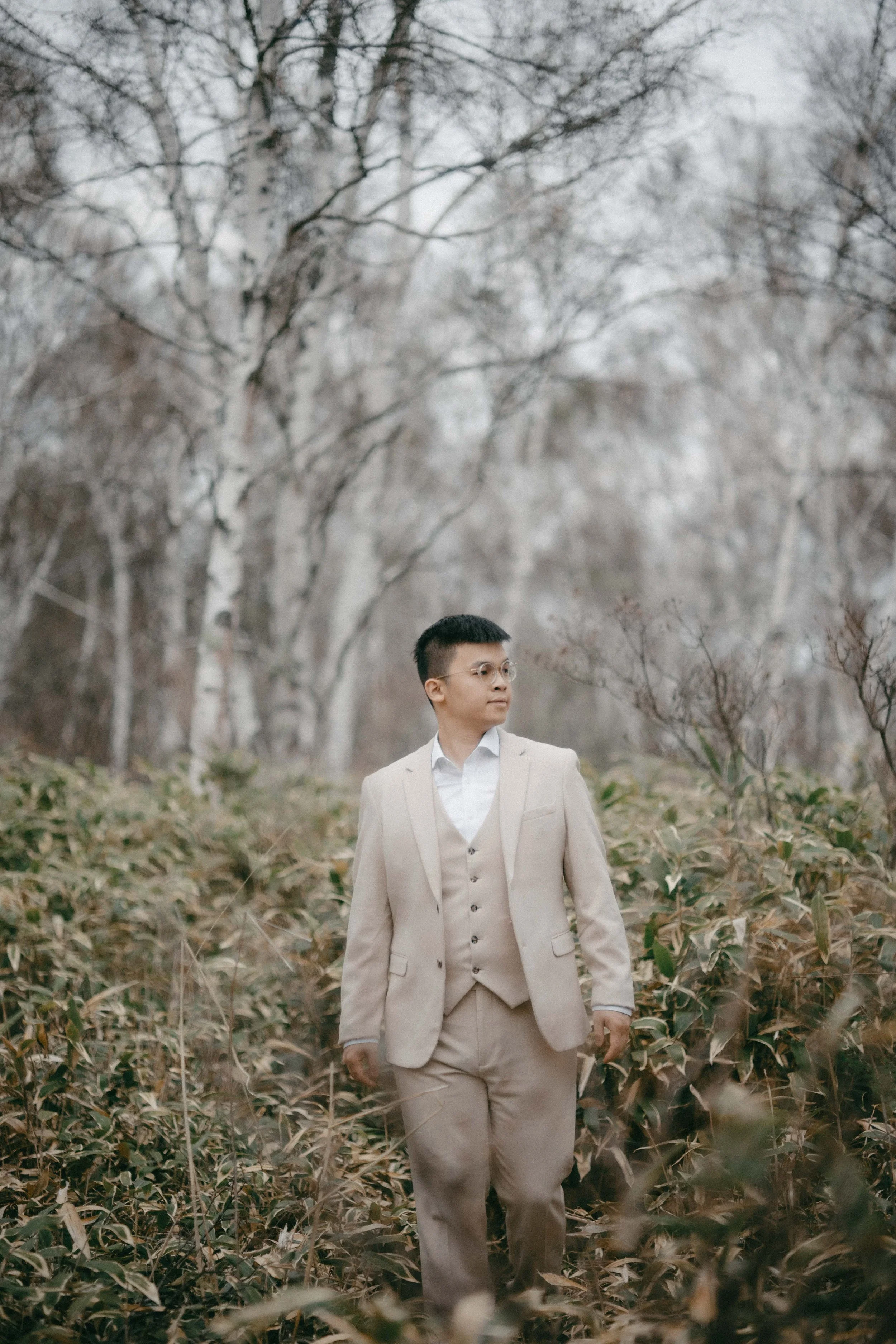 A man in a beige suit and glasses walking through a forest with leafless trees and brown foliage.