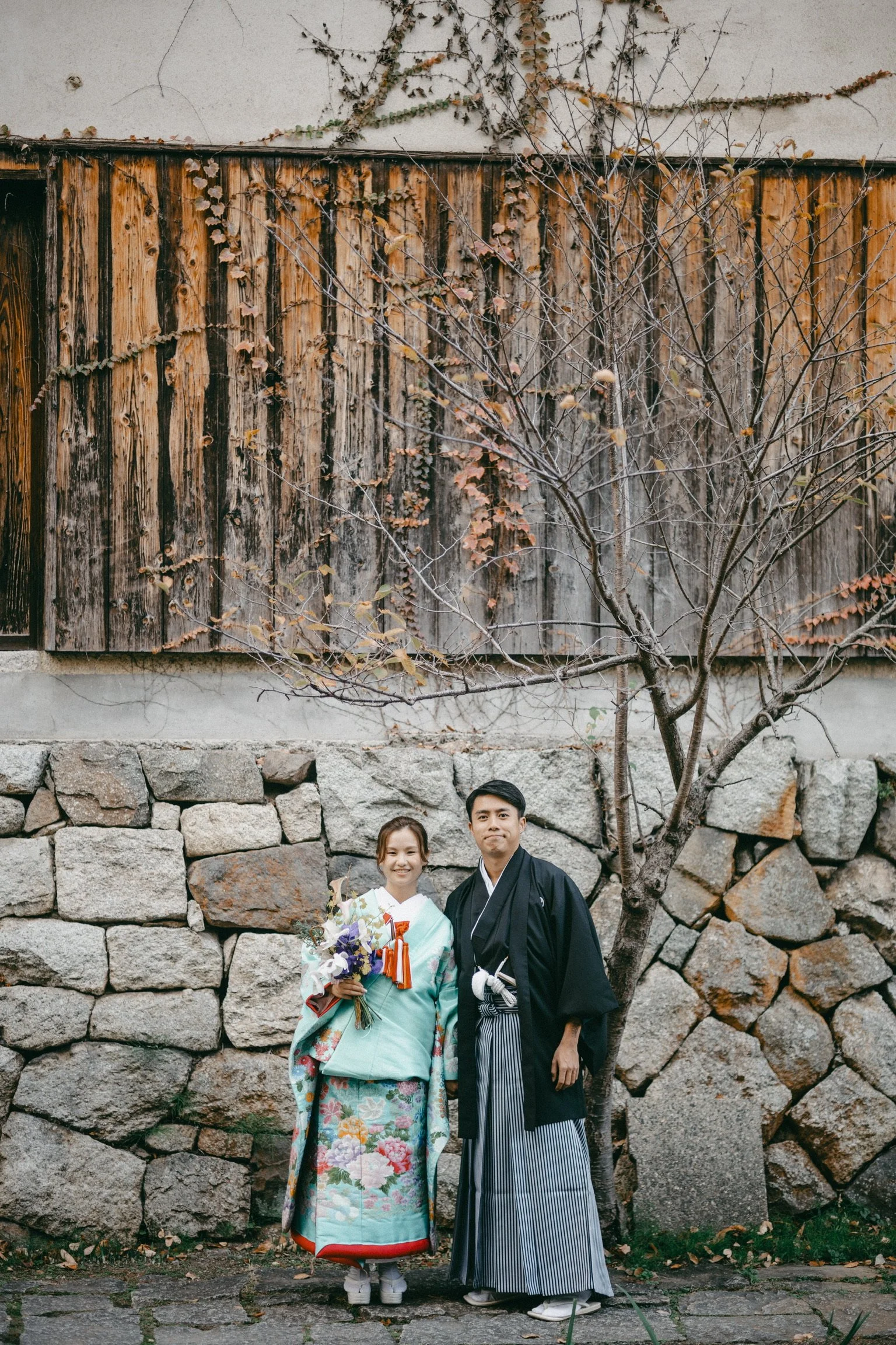 A young woman and a young man in traditional Japanese kimonos and hakamas standing outdoors in front of a stone wall and a leafless tree, holding a bouquet of flowers.