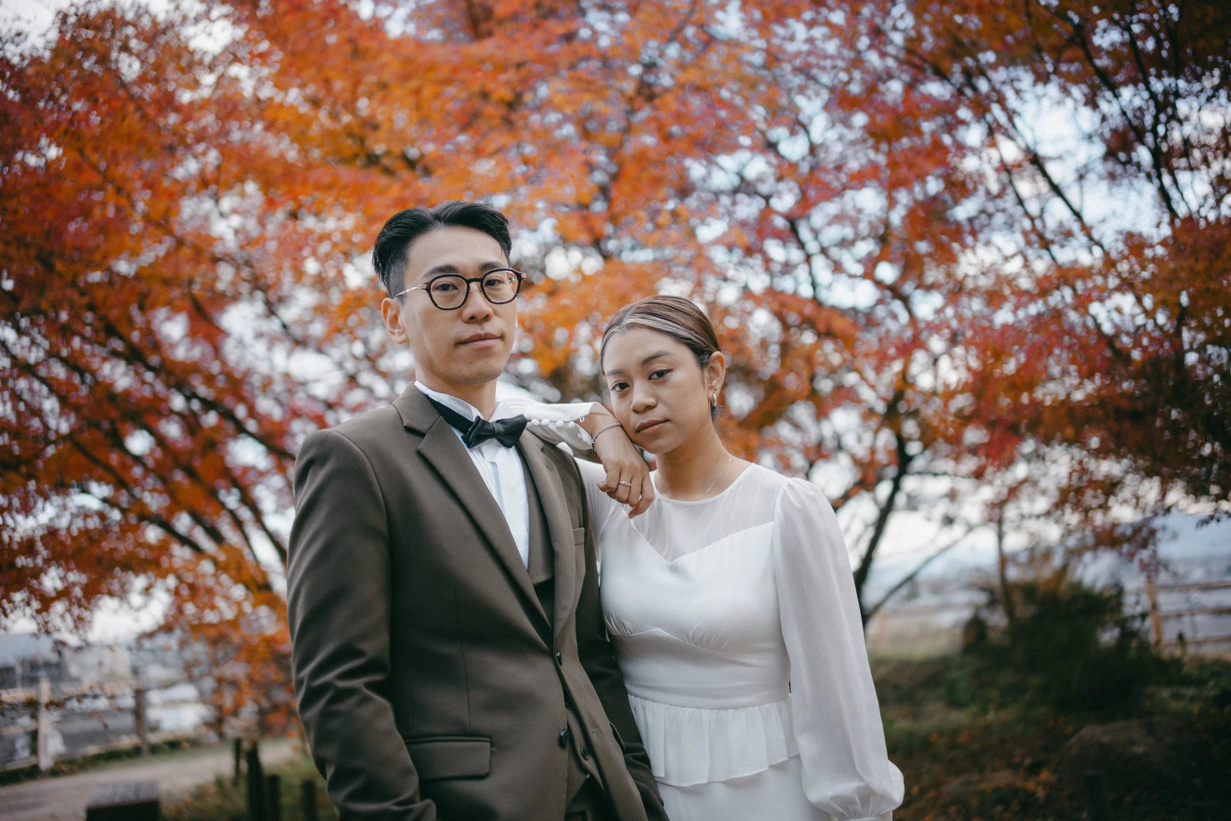 A young couple standing outdoors in front of a tree with vibrant orange and red fall foliage. The man is wearing glasses, a brown suit with a bow tie, and a white shirt. The woman is dressed in a white dress with long sleeves. The woman has her arm r