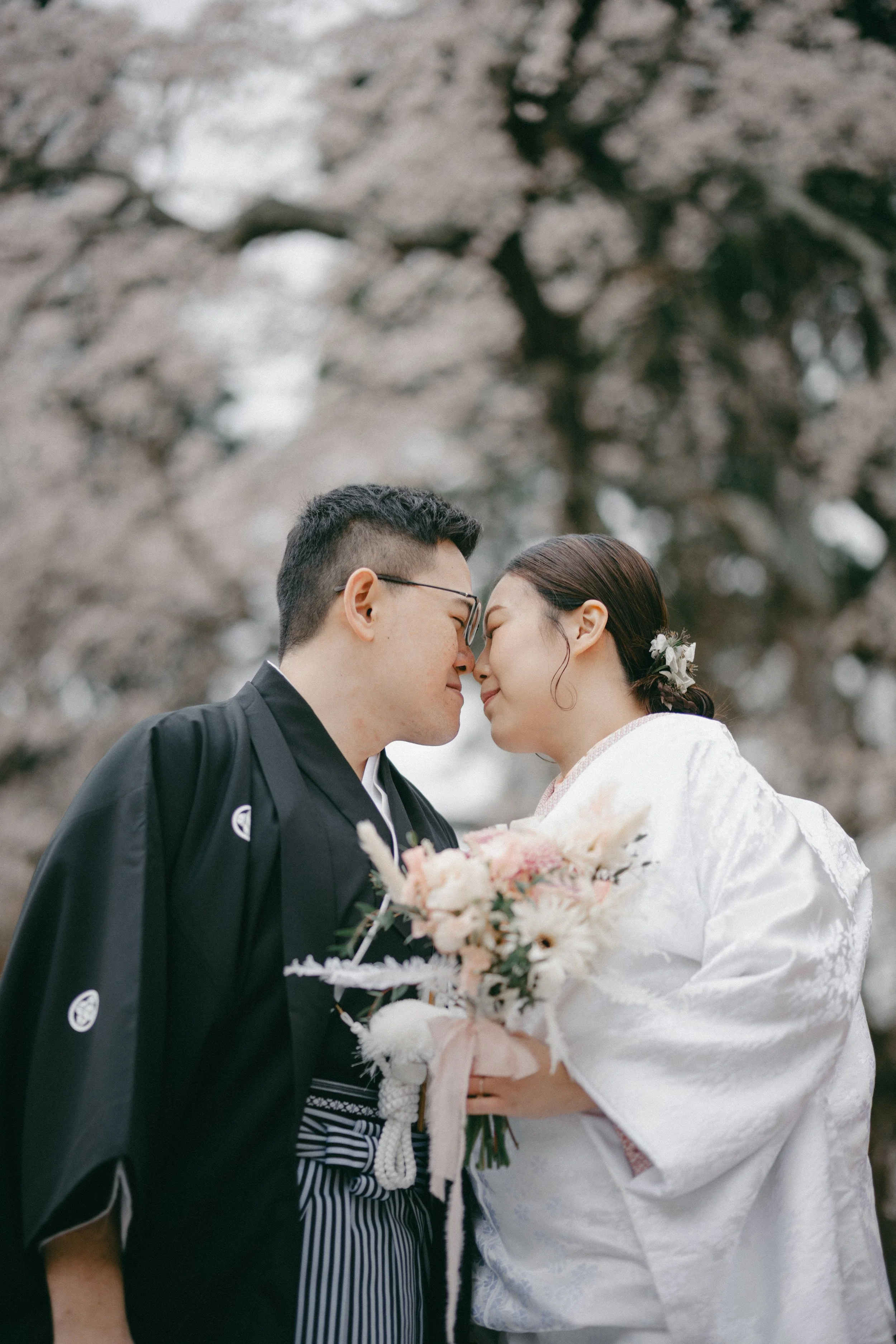 A couple dressed in traditional Japanese wedding attire embracing with foreheads touching, holding a bouquet of flowers, surrounded by cherry blossom trees.
