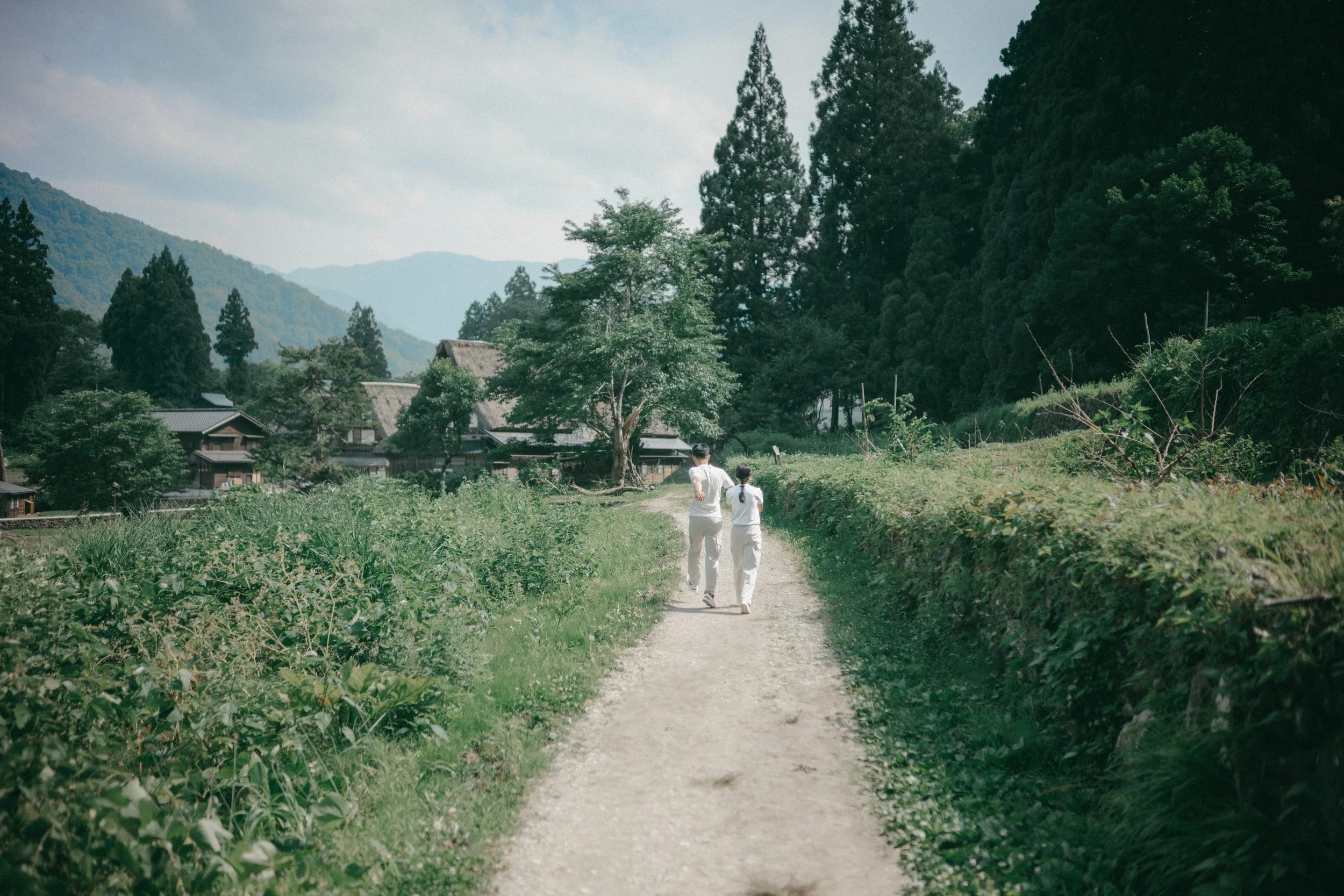 Two people walking on a dirt path through a rural area with green fields, trees, traditional houses, and mountains in the background.