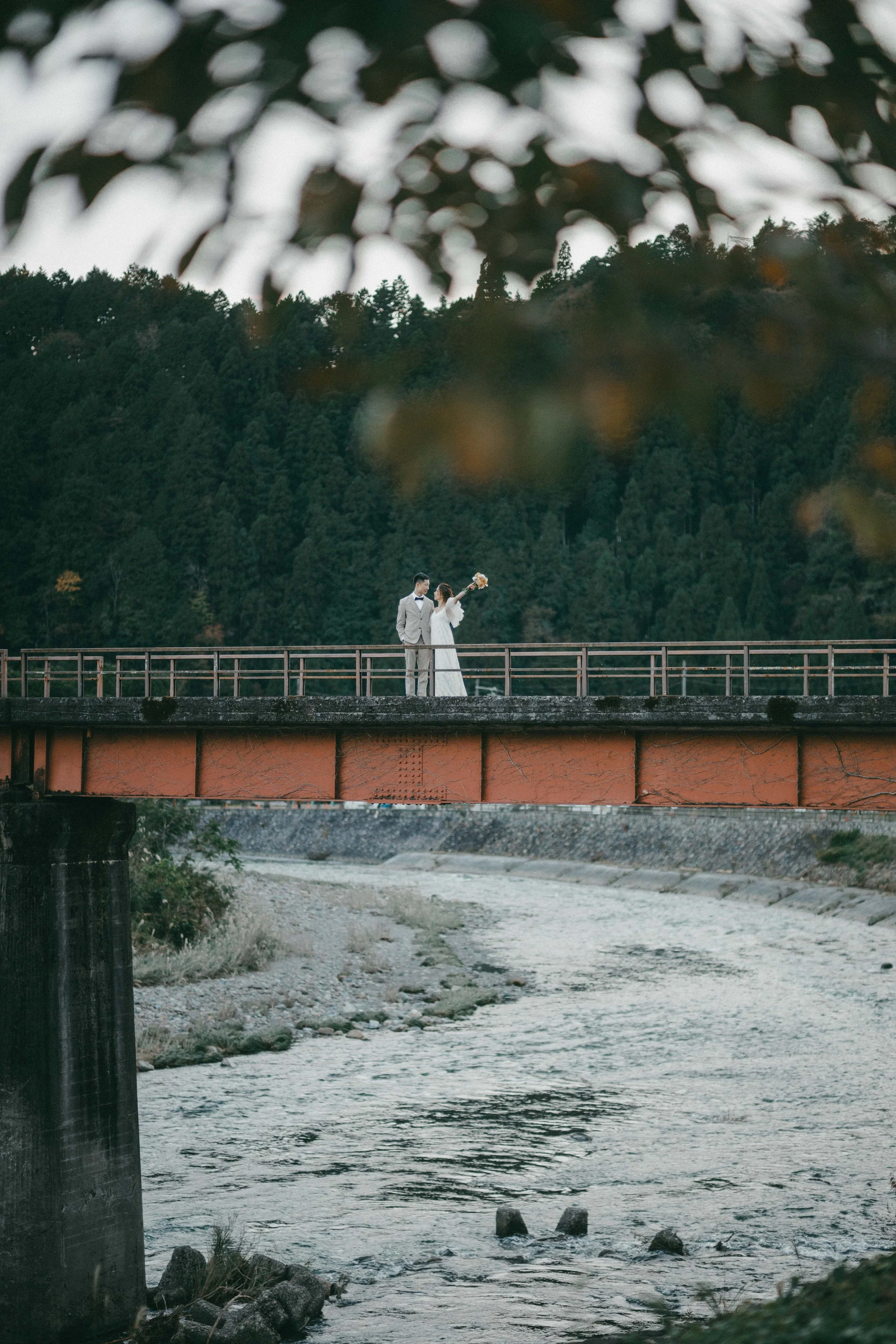 A bride and groom standing on a bridge over a river with a forested mountain background.
