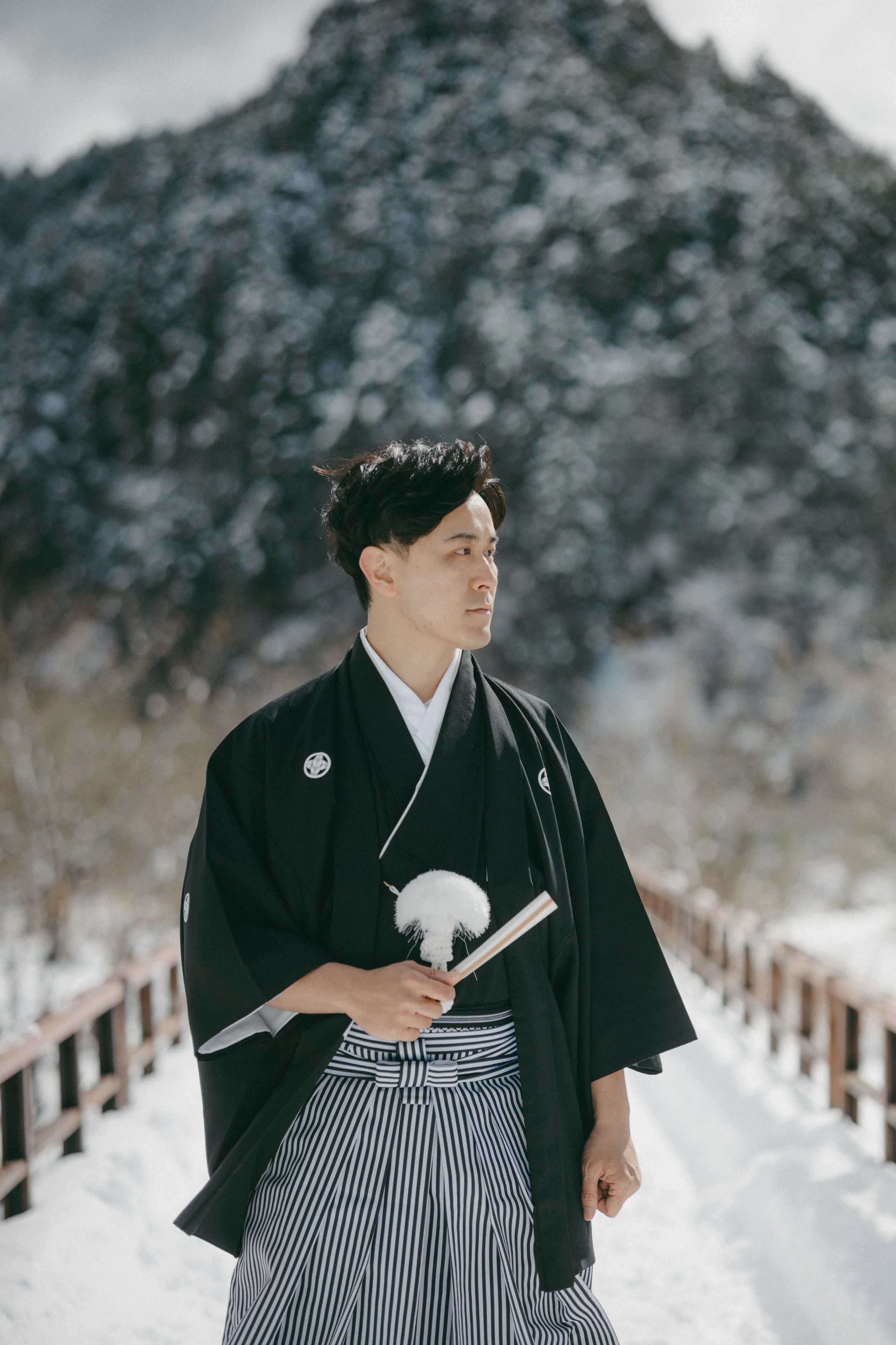 A man dressed in traditional Japanese kimono, holding a fan, standing on a snow-covered bridge with a mountain in the background.