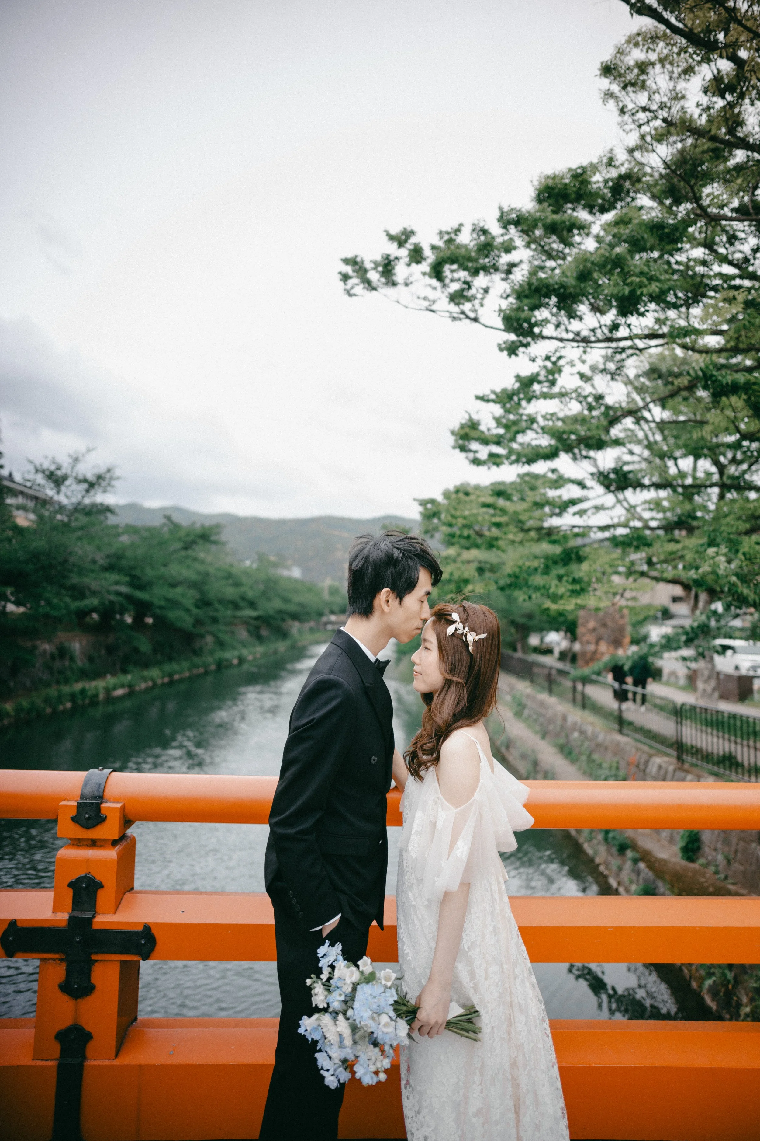 A bride and groom standing close on a bridge over a river, with trees and mountains in the background. The groom in a black tuxedo is leaning down to touch foreheads with the bride, who is wearing a white lace wedding dress and holding a bouquet of w