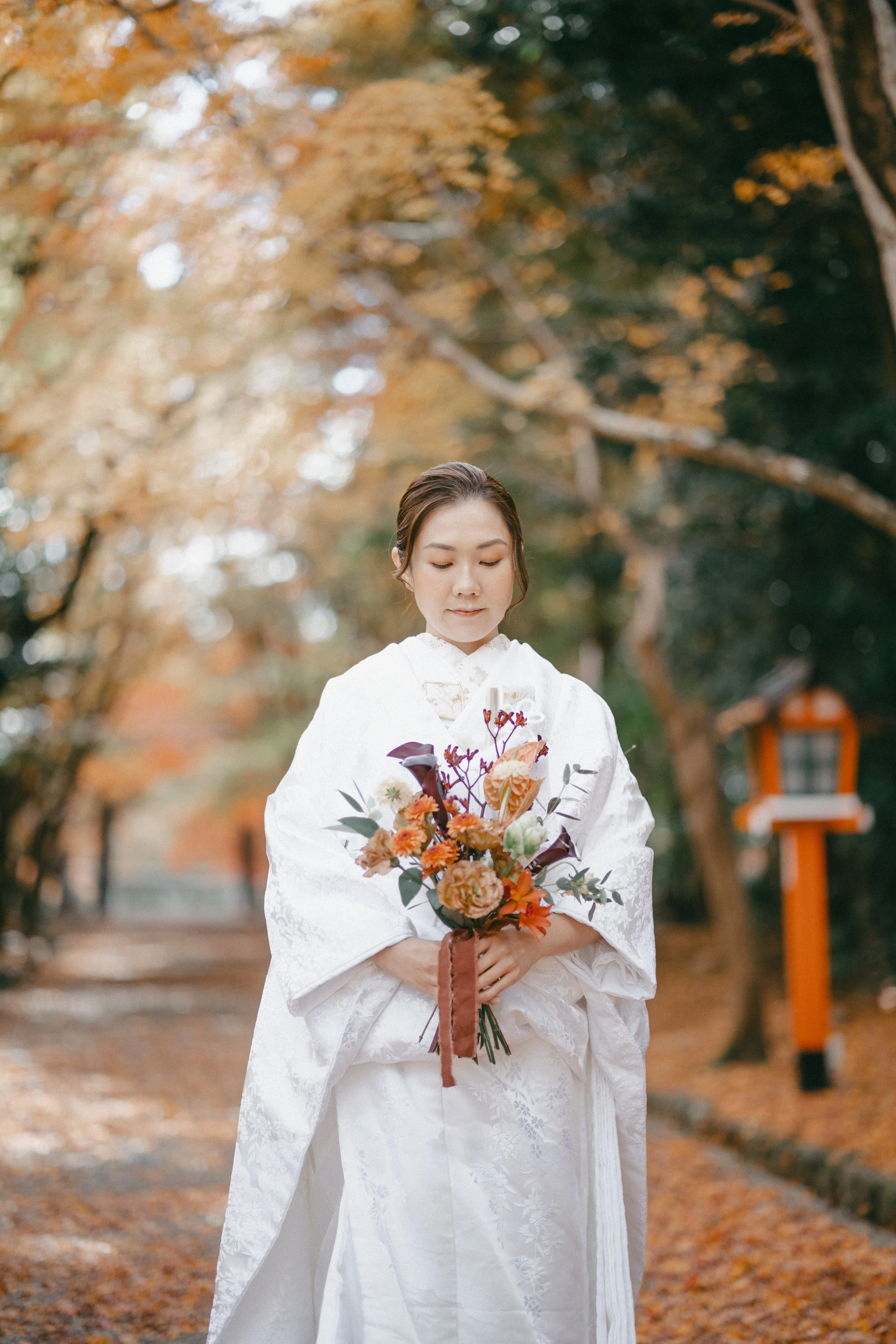 A woman in traditional white Japanese wedding kimono holding a colorful bouquet of flowers in an outdoor setting with autumn leaves.