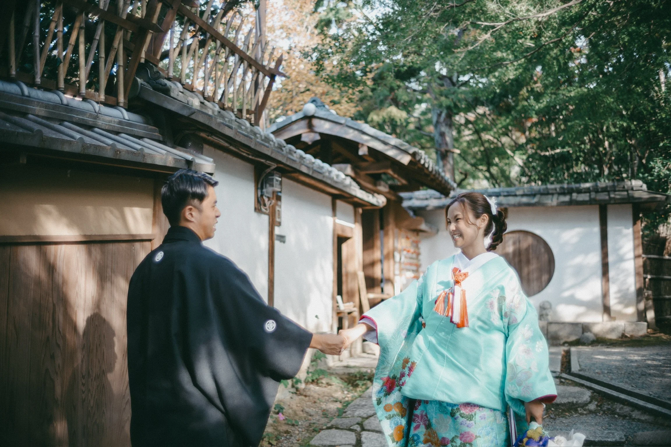 A young woman in a pastel teal kimono with floral patterns and a bright smile shaking hands with a man in a traditional black kimono. They are outdoors in front of a traditional Japanese building with tiled roofs and wooden sliding doors, surrounded 