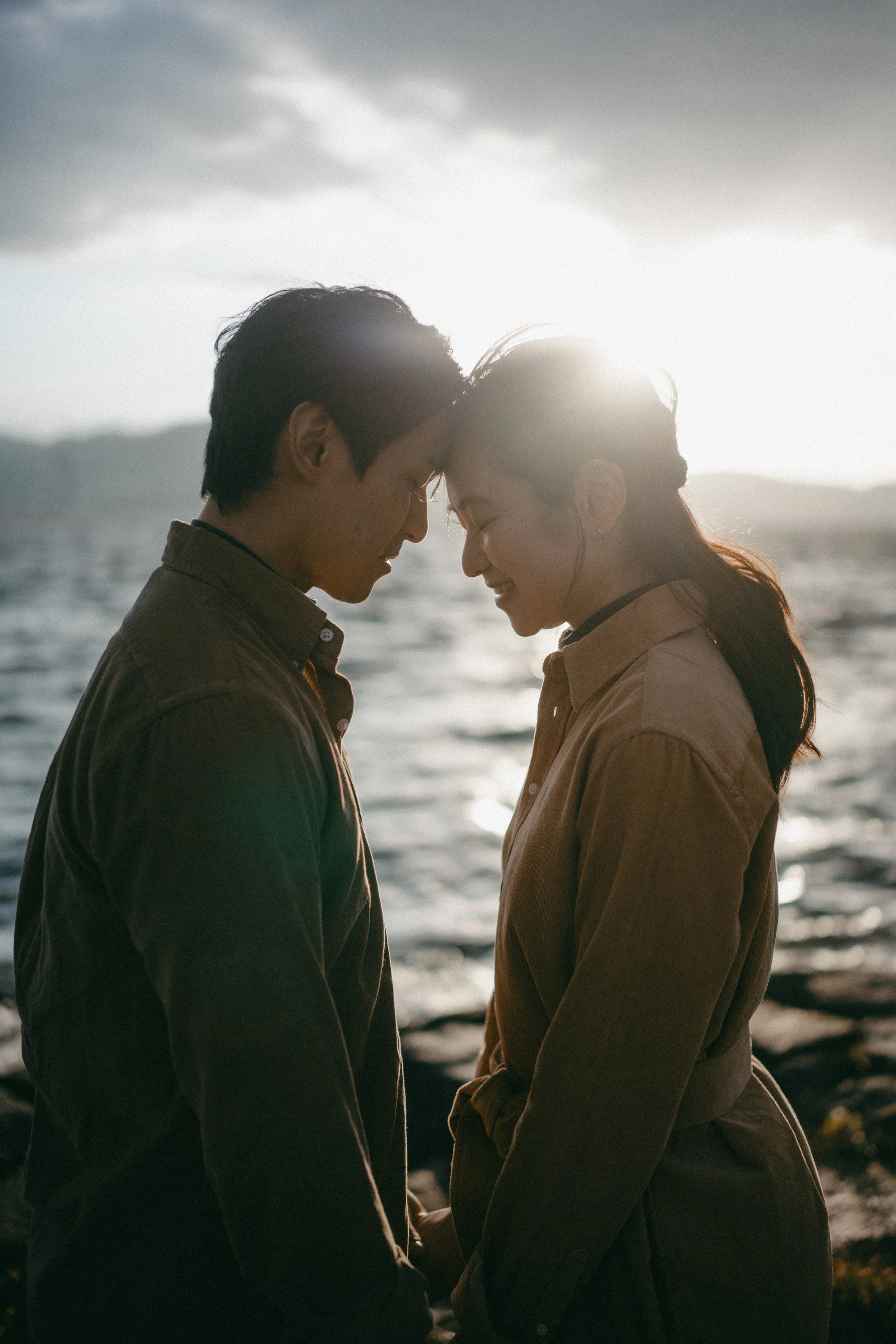 A couple standing close together on the shoreline with their foreheads touching, backlit by the setting sun, near the ocean with waves and rocks.