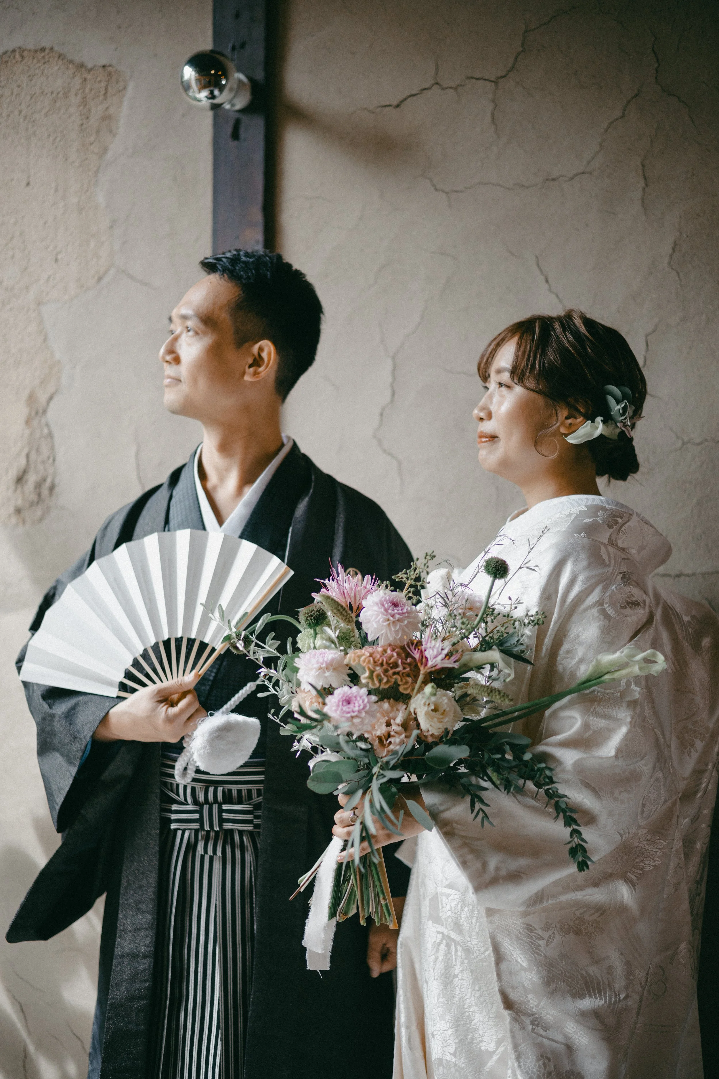 A couple dressed in traditional Japanese wedding attire, standing side by side, with the woman holding a bouquet and the man holding a fan, against a rustic wall.
