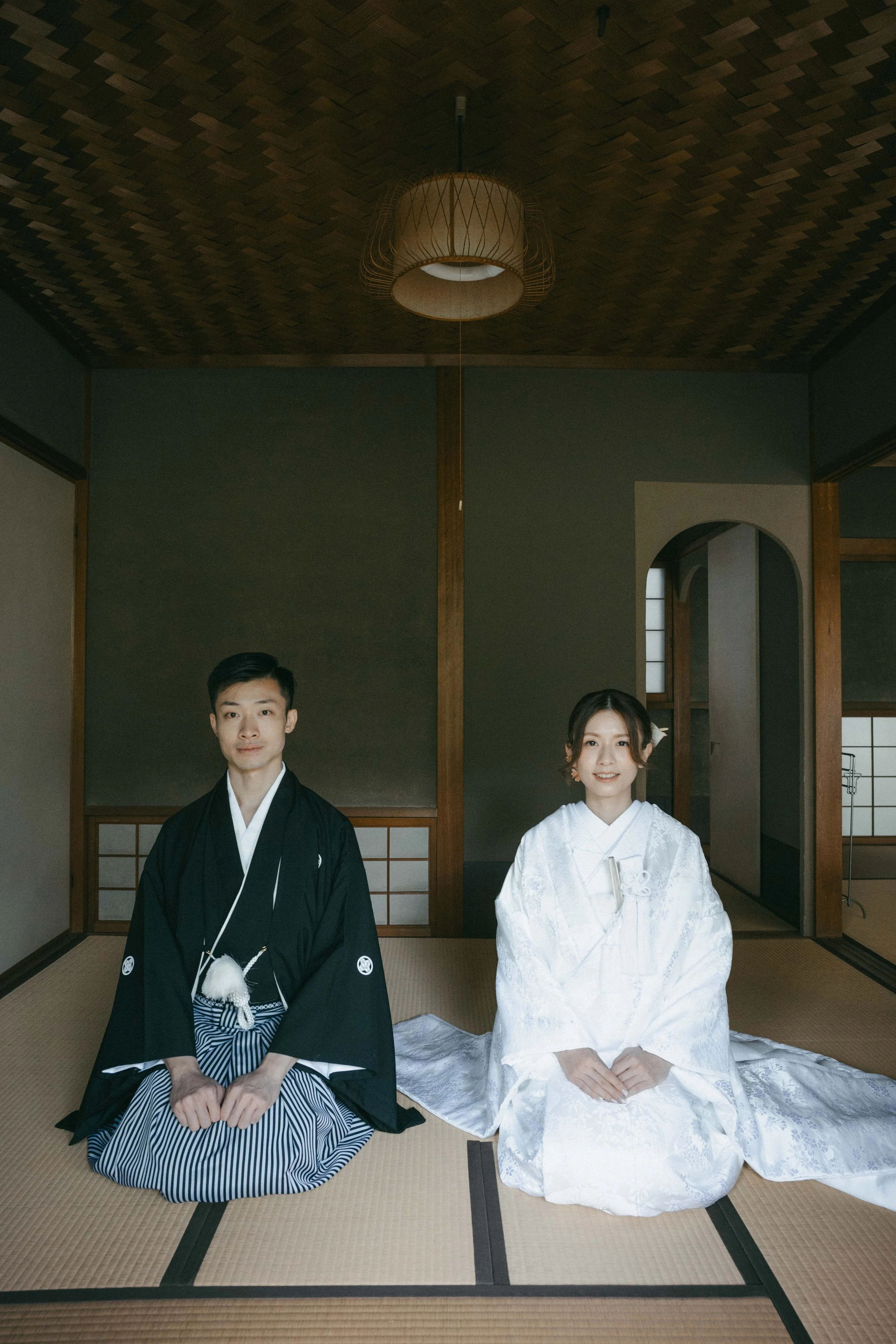 A couple dressed in traditional Japanese wedding attire, sitting on tatami mats in a Japanese-style room.