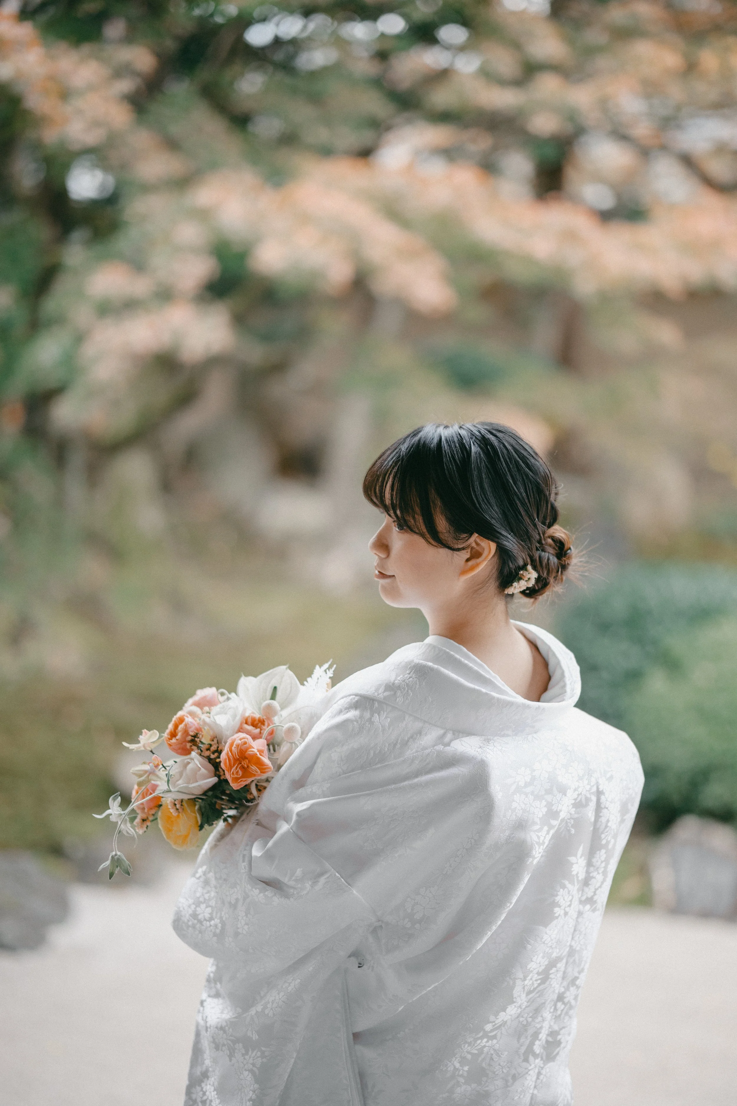 A woman in a white traditional kimono holding a bouquet of flowers, standing outdoors with autumn foliage in the background.