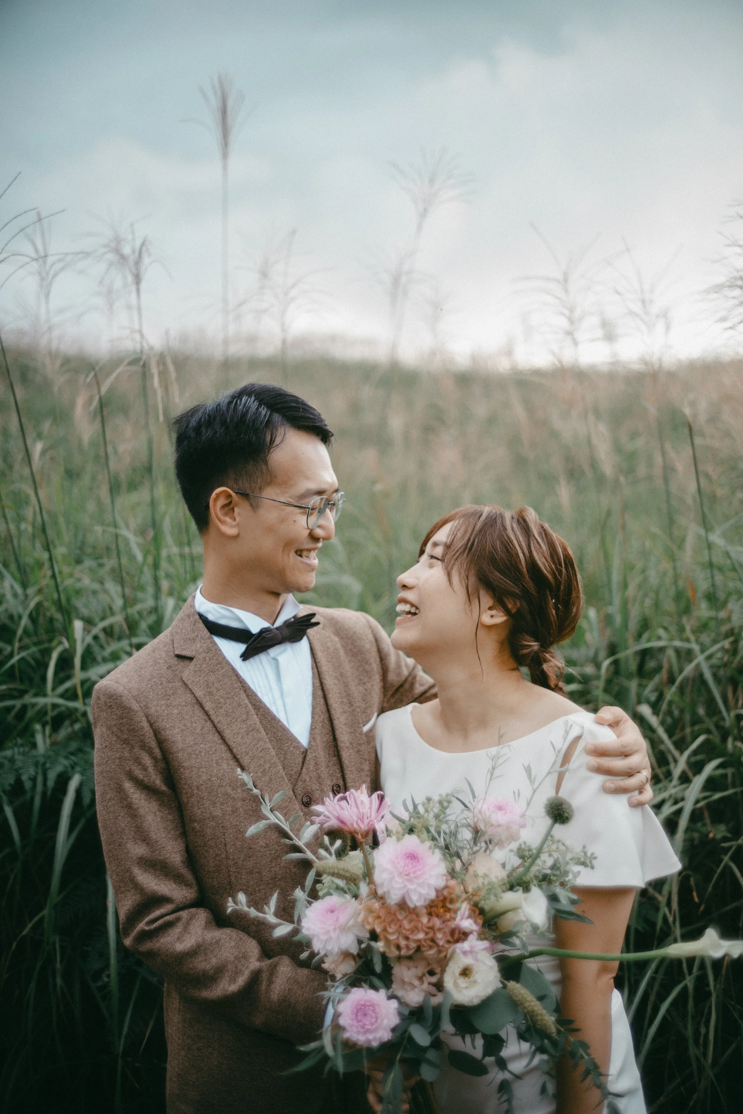 A happy couple on their wedding day, the groom wearing glasses, a brown suit and bow tie, the bride in a white dress holding a large bouquet of flowers, standing in a field of tall grass.