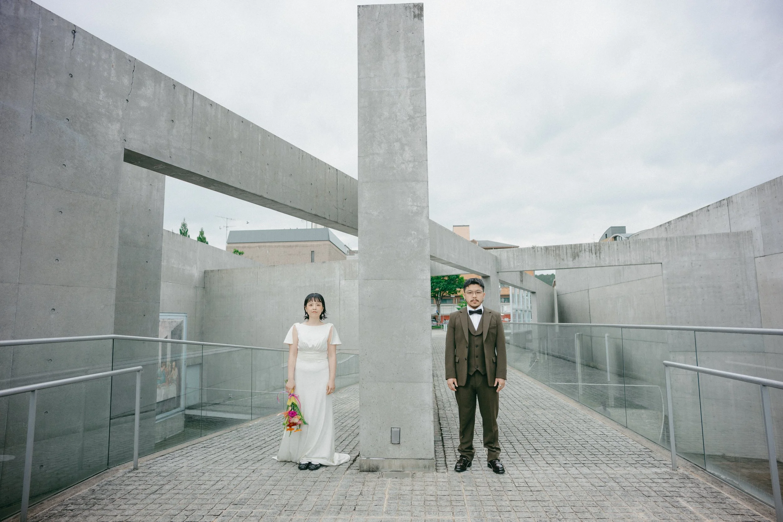 A man and a woman stand apart on a paved walkway, separated by a large concrete structure, in an urban outdoor setting. The woman wears a white dress and holds a colorful bouquet, while the man is dressed in a dark suit with a bow tie.