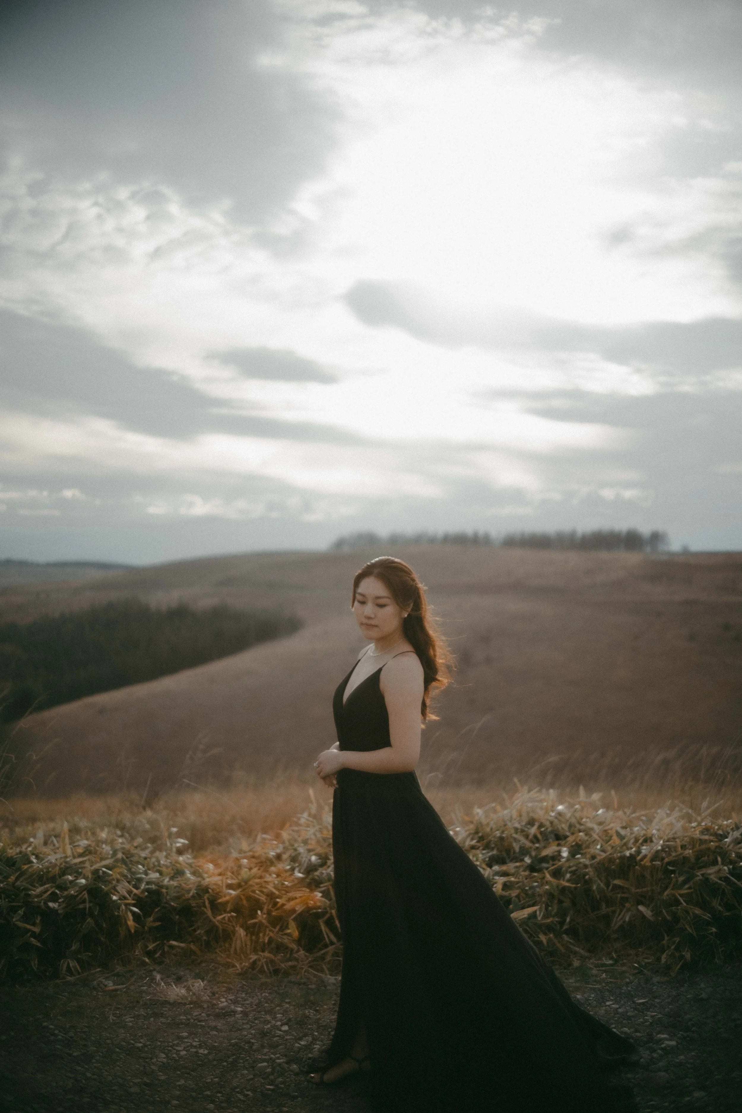 A woman in a long black dress standing outdoors in a field with rolling hills and a cloudy sky in the background.