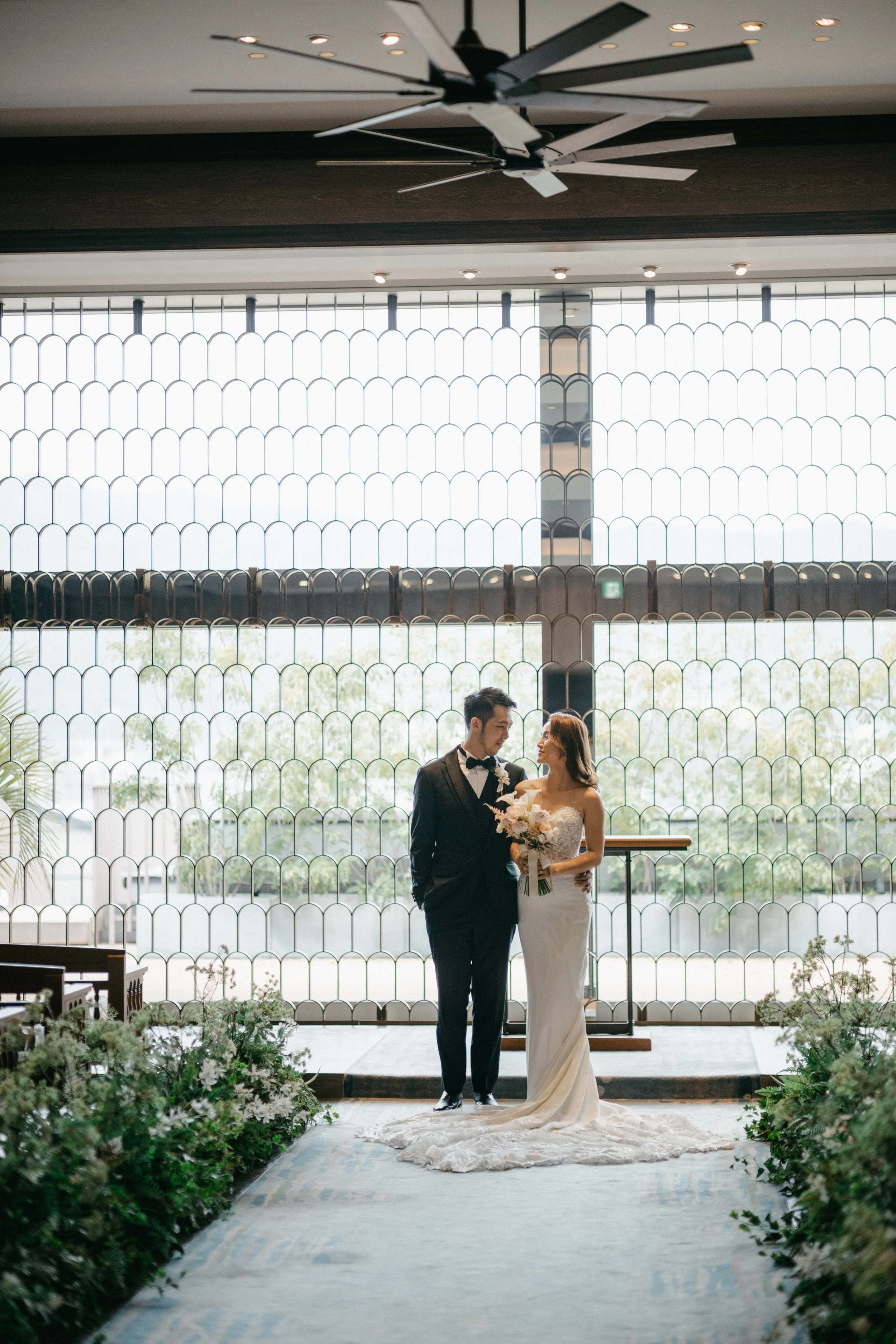 A bride and groom standing together in front of a large window with a vintage stained glass pattern. The bride is holding a bouquet, and both are dressed in wedding attire. The scene is decorated with greenery and flowers along the aisle.