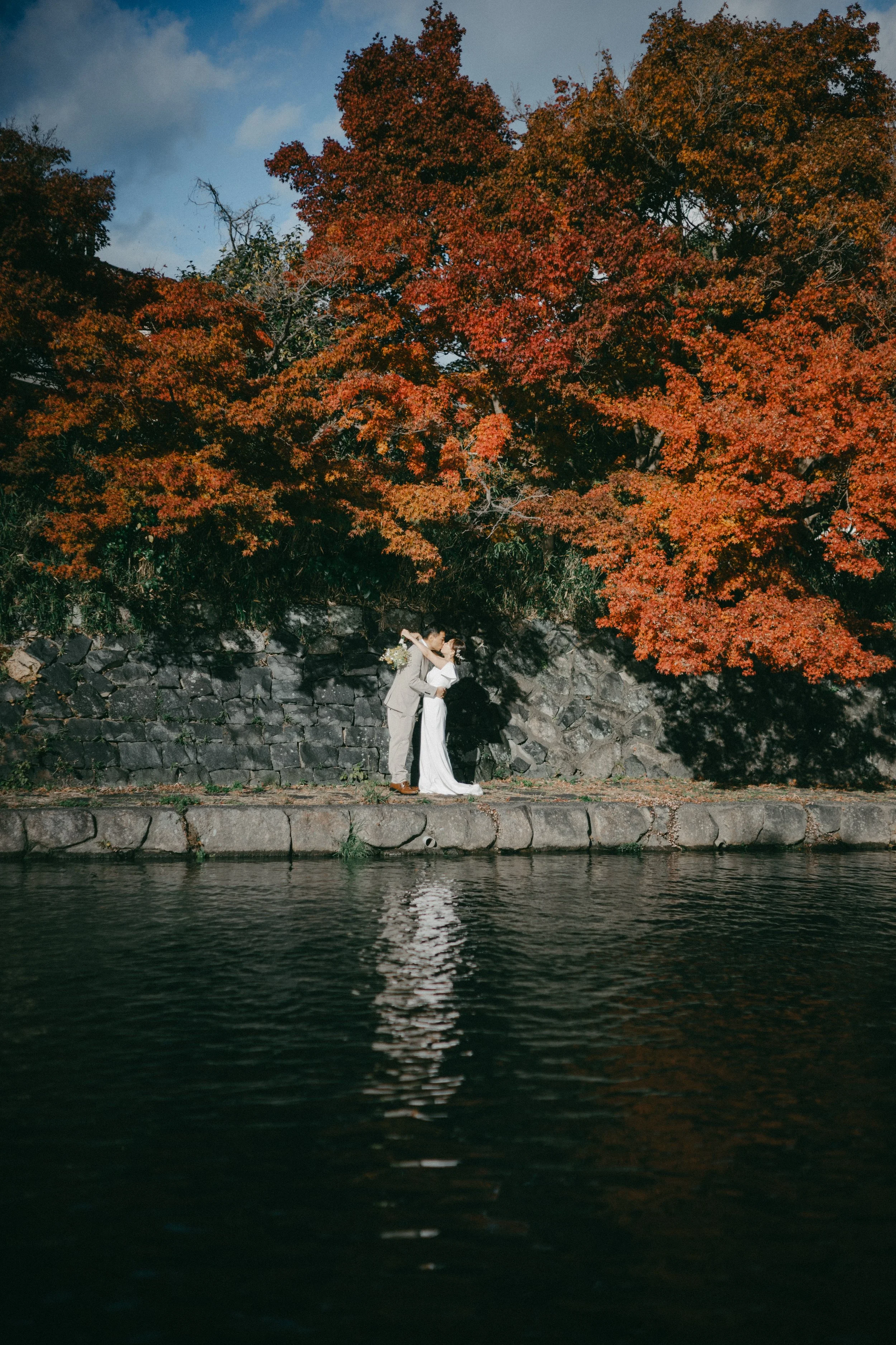A couple in wedding attire standing on a stone path beside a body of water with autumn-colored trees in the background.