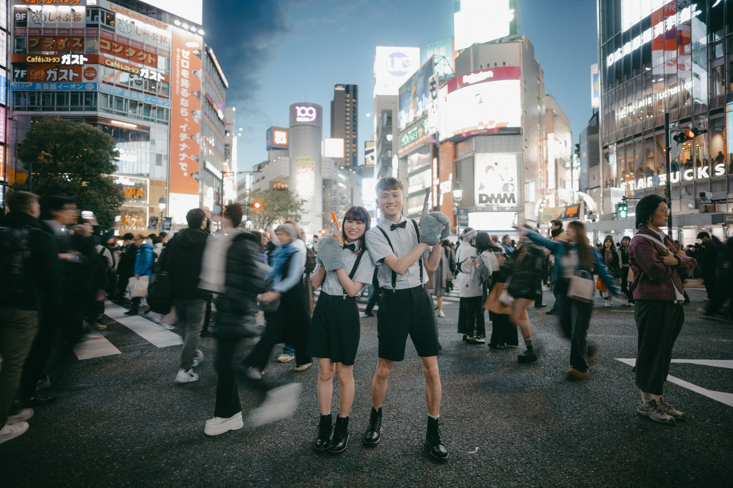 two young people dressed as waiters with gray gloves and bow ties standing in the middle of a busy city street at night, surrounded by pedestrians and bright digital billboards.