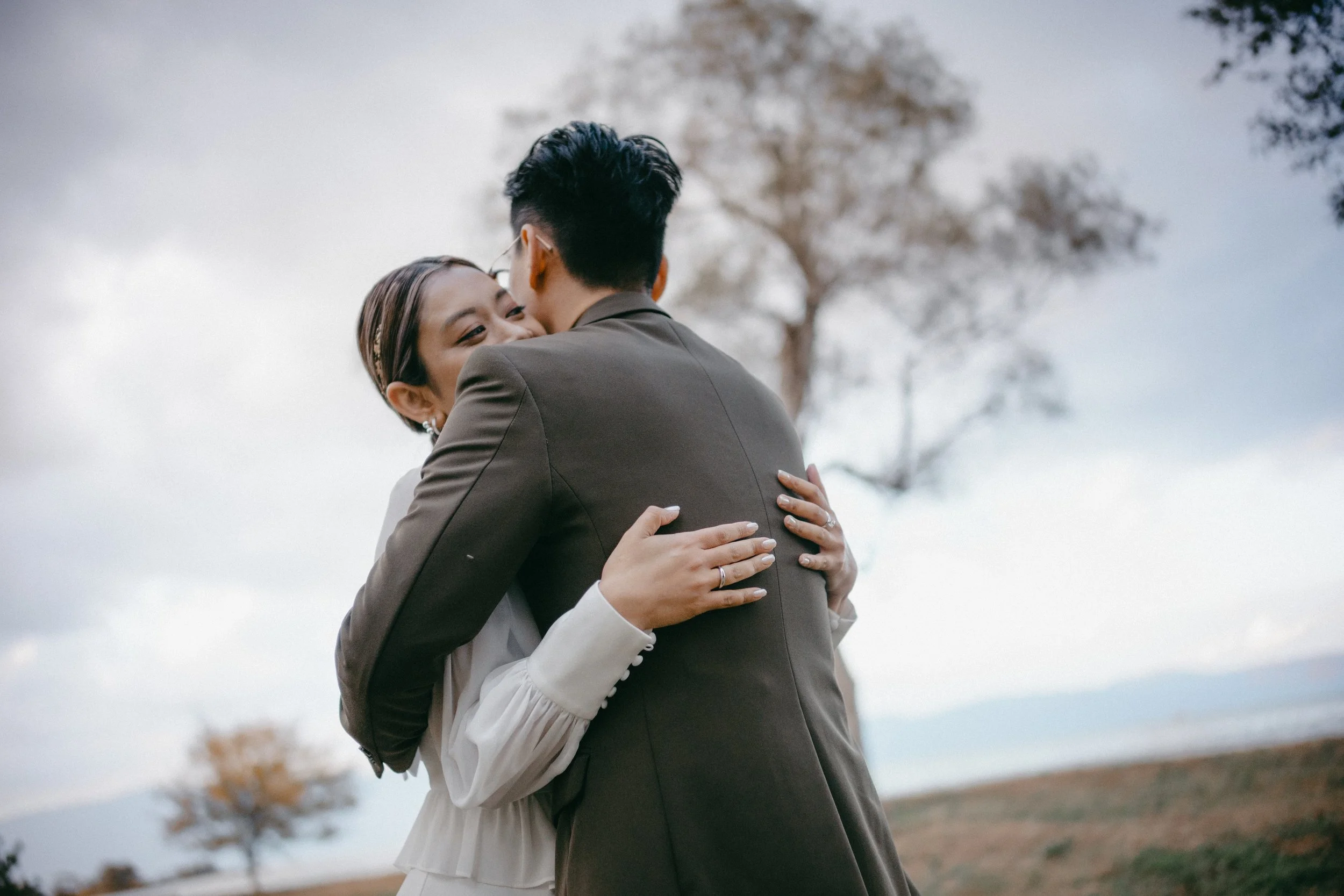 A couple hugging outdoors with trees and cloudy sky in the background.