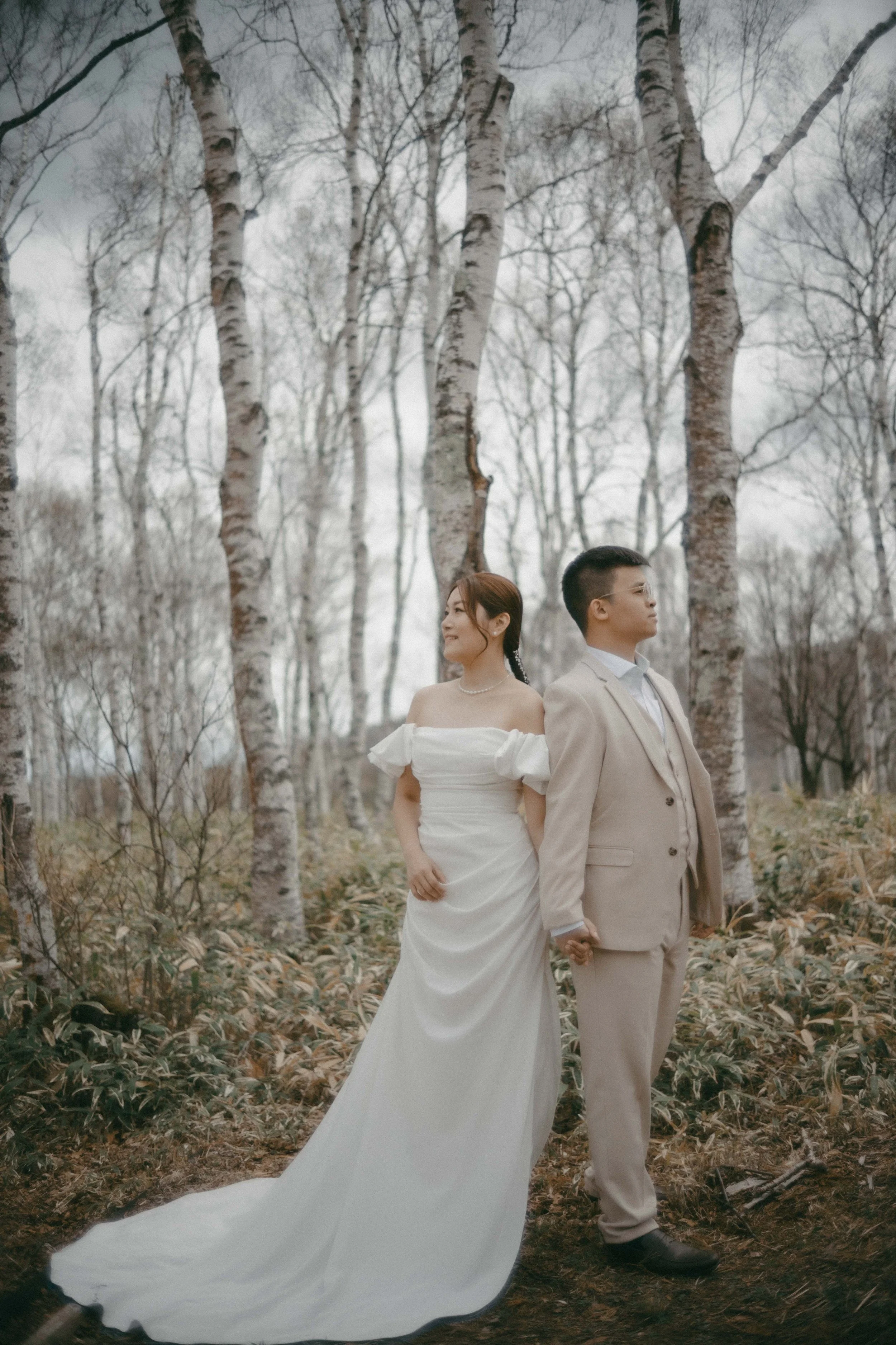 A couple dressed in wedding attire standing hand in hand in a forest of bare trees, with the woman in a white off-the-shoulder wedding gown and the man in a light-colored suit.