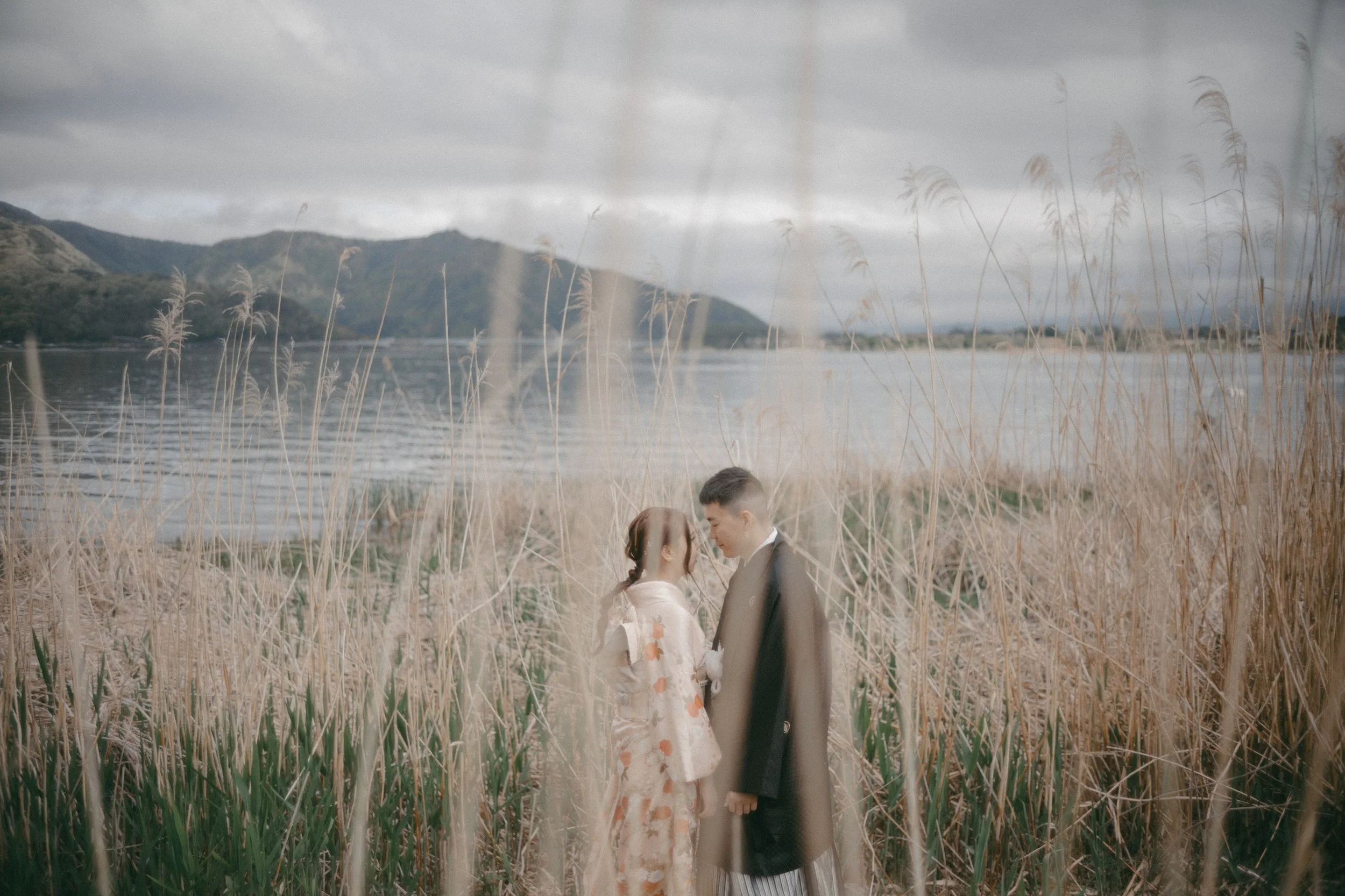 A couple dressed in traditional Japanese attire standing close together in a field of tall dry grasses by a body of water with mountains in the background, under a cloudy sky.