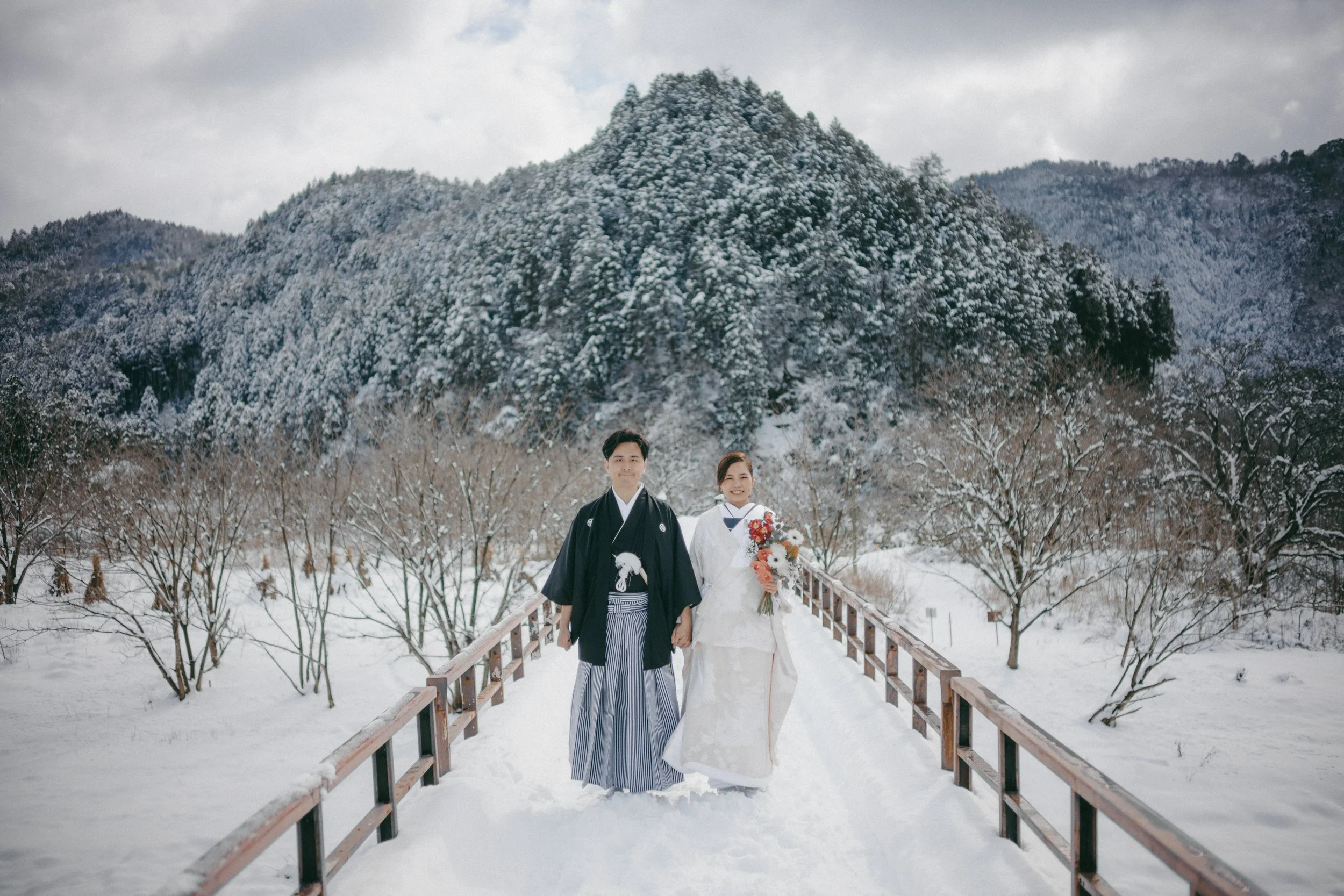 A couple in traditional Japanese wedding attire standing hand in hand on a snowy path with a snow-covered mountain in the background.