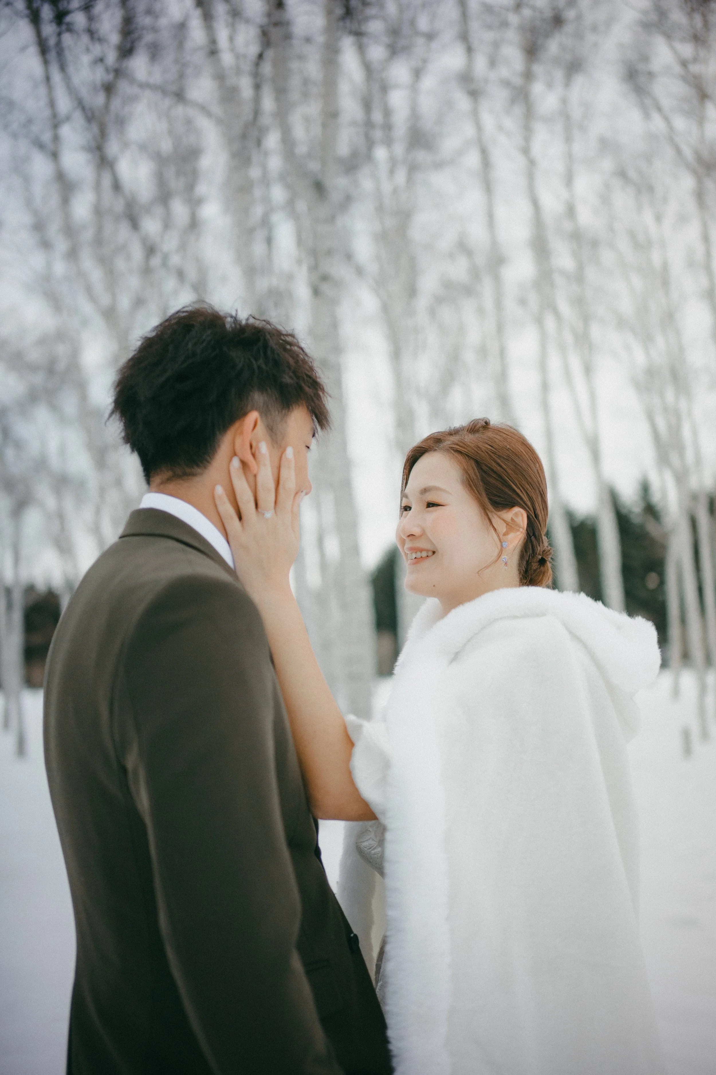 A couple standing outdoors in winter, the woman holding the man's face and smiling at him, snow-covered trees in the background.