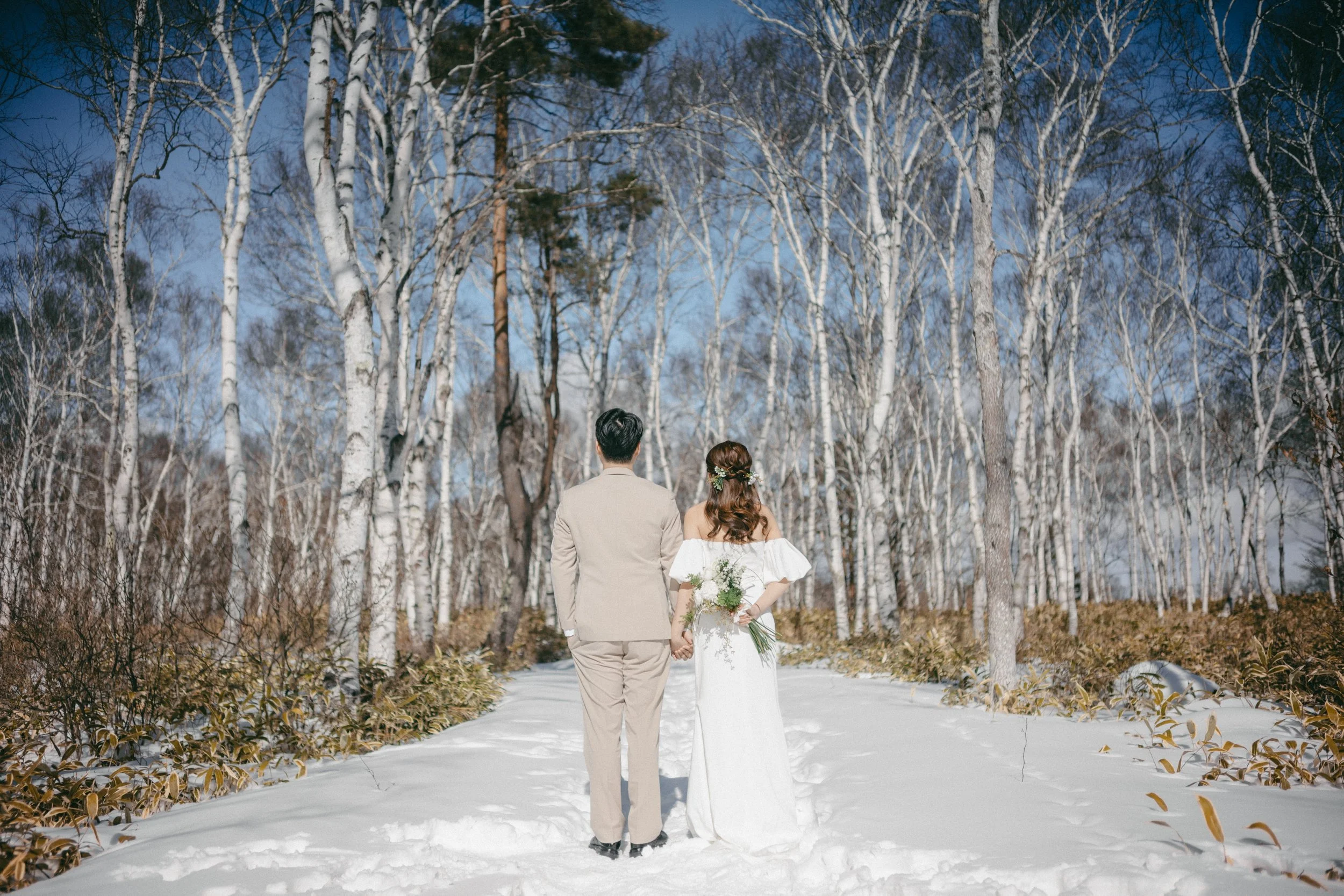 A couple dressed in wedding attire walking hand in hand through a snowy forest with bare trees and a blue sky.