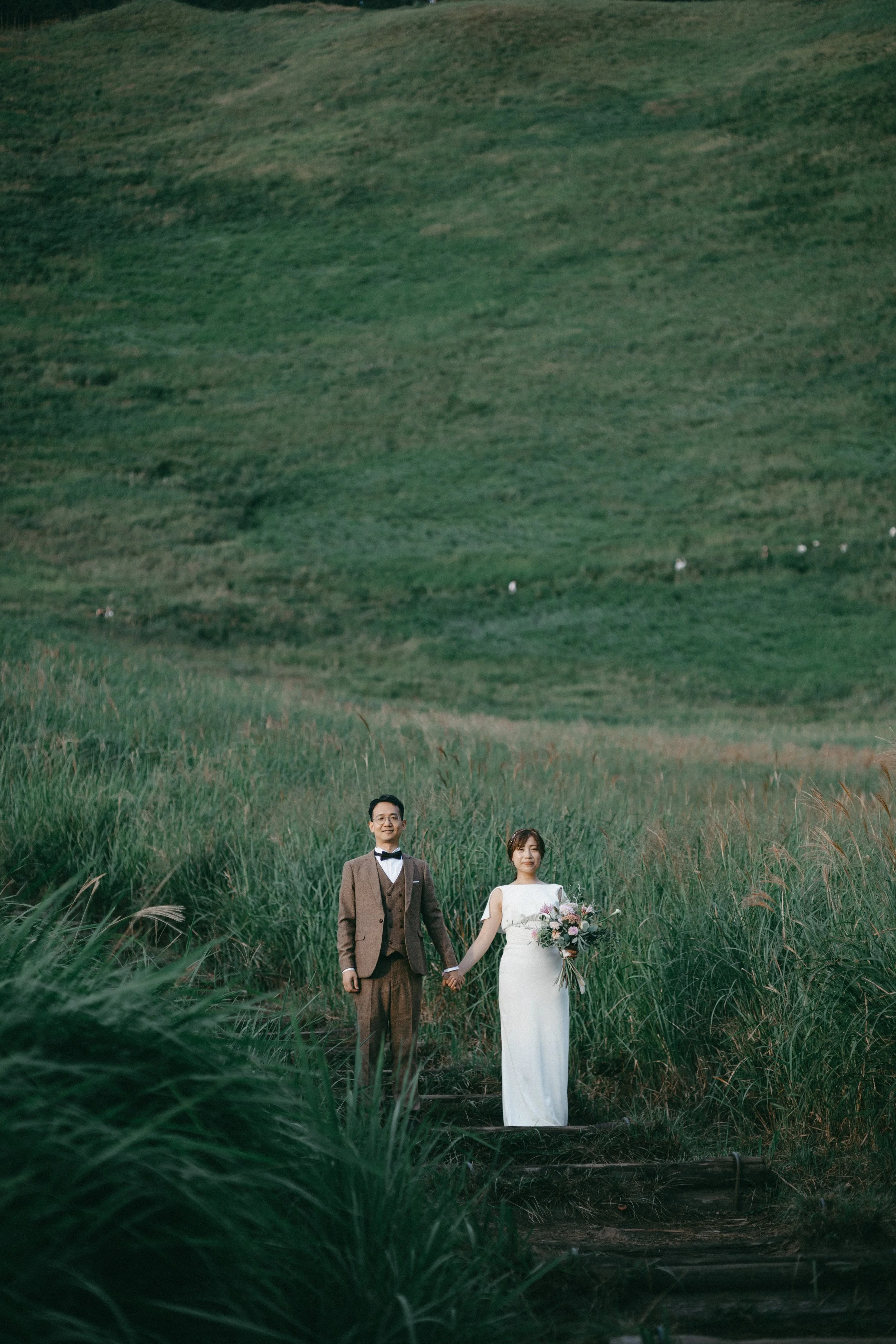 A bride and groom holding hands stand in a lush, green field with rolling hills in the background. The bride is in a white wedding dress holding a bouquet, and the groom is in a brown suit with a bow tie.