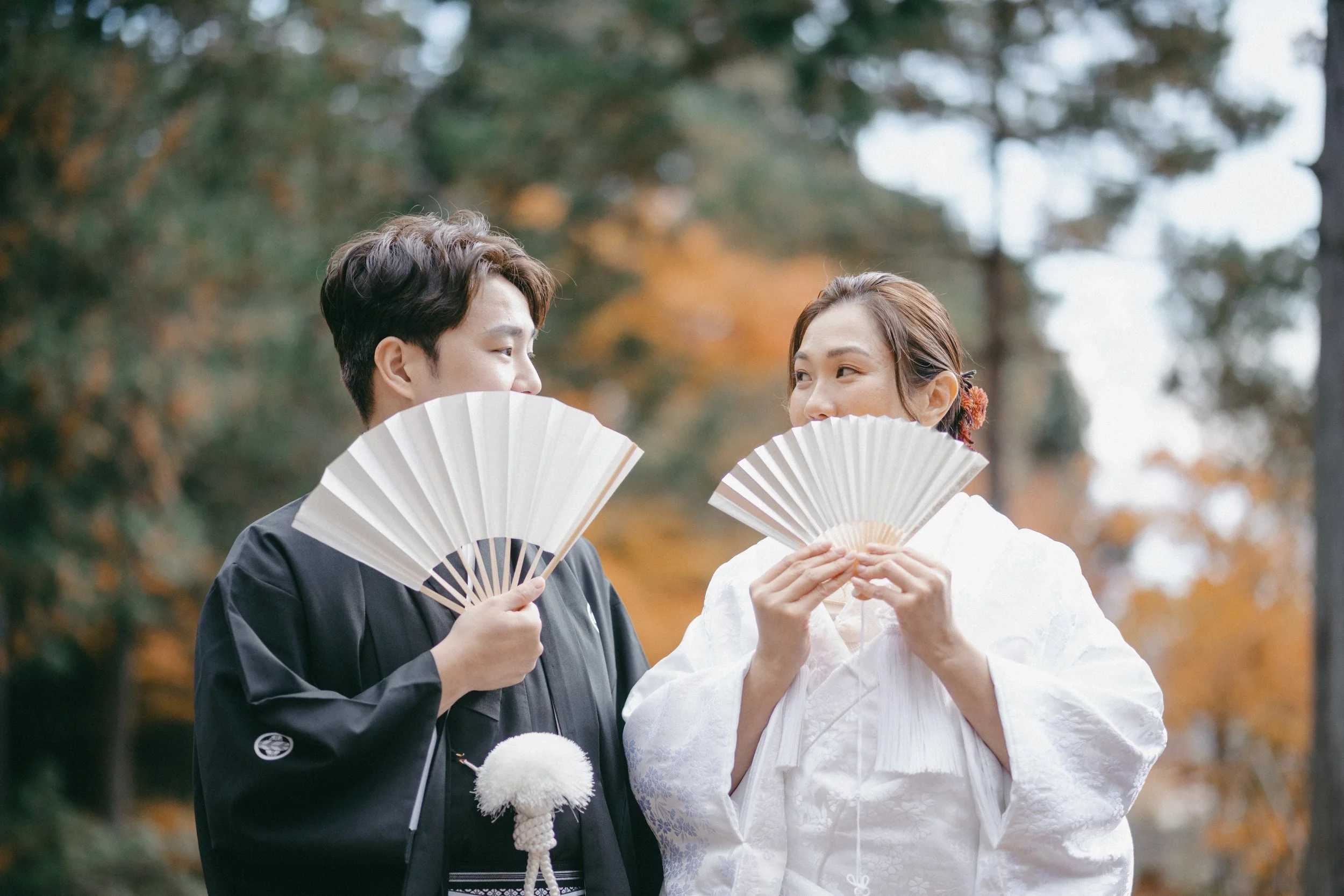 A young man and woman dressed in traditional Japanese wedding attire, holding white folding fans, standing outdoors with autumn-colored trees in the background, looking at each other.