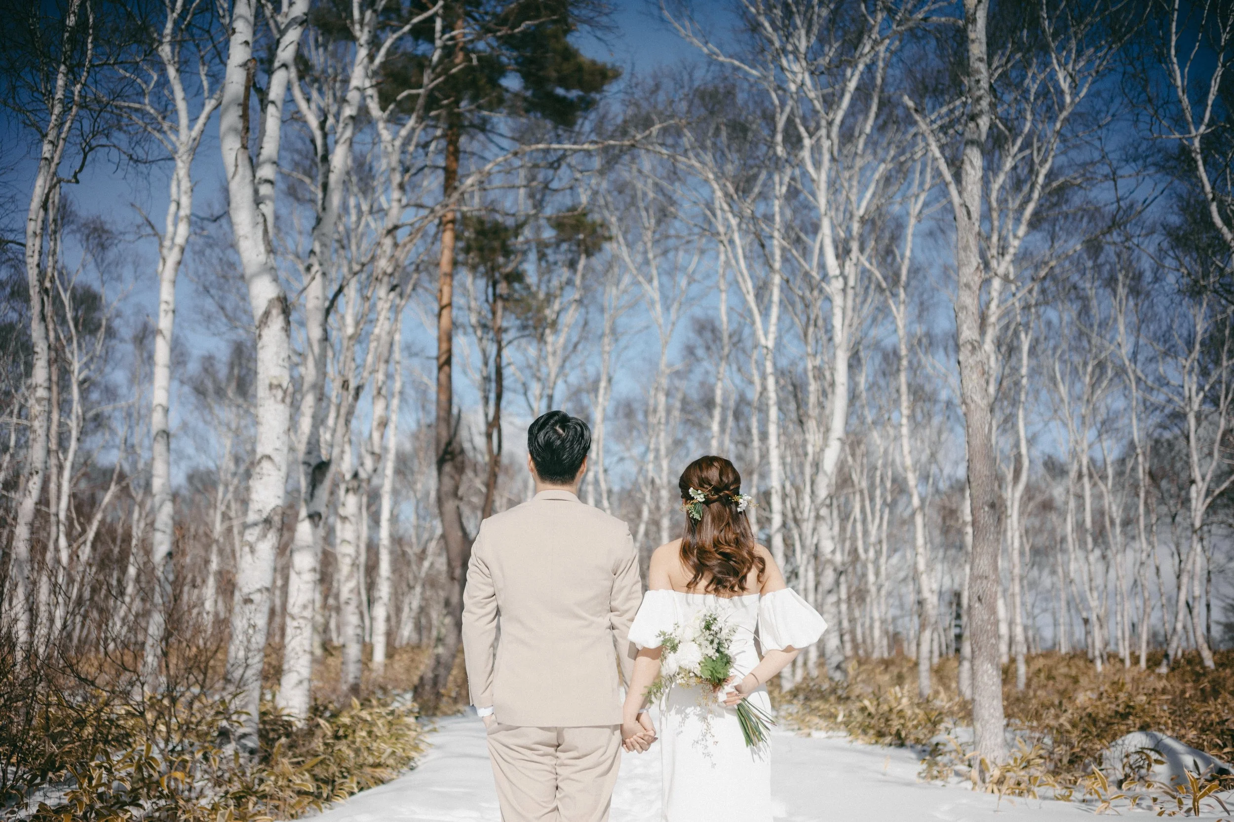 A couple dressed in wedding attire holding hands and walking away in a snow-covered forest with tall, leafless trees under a clear blue sky.