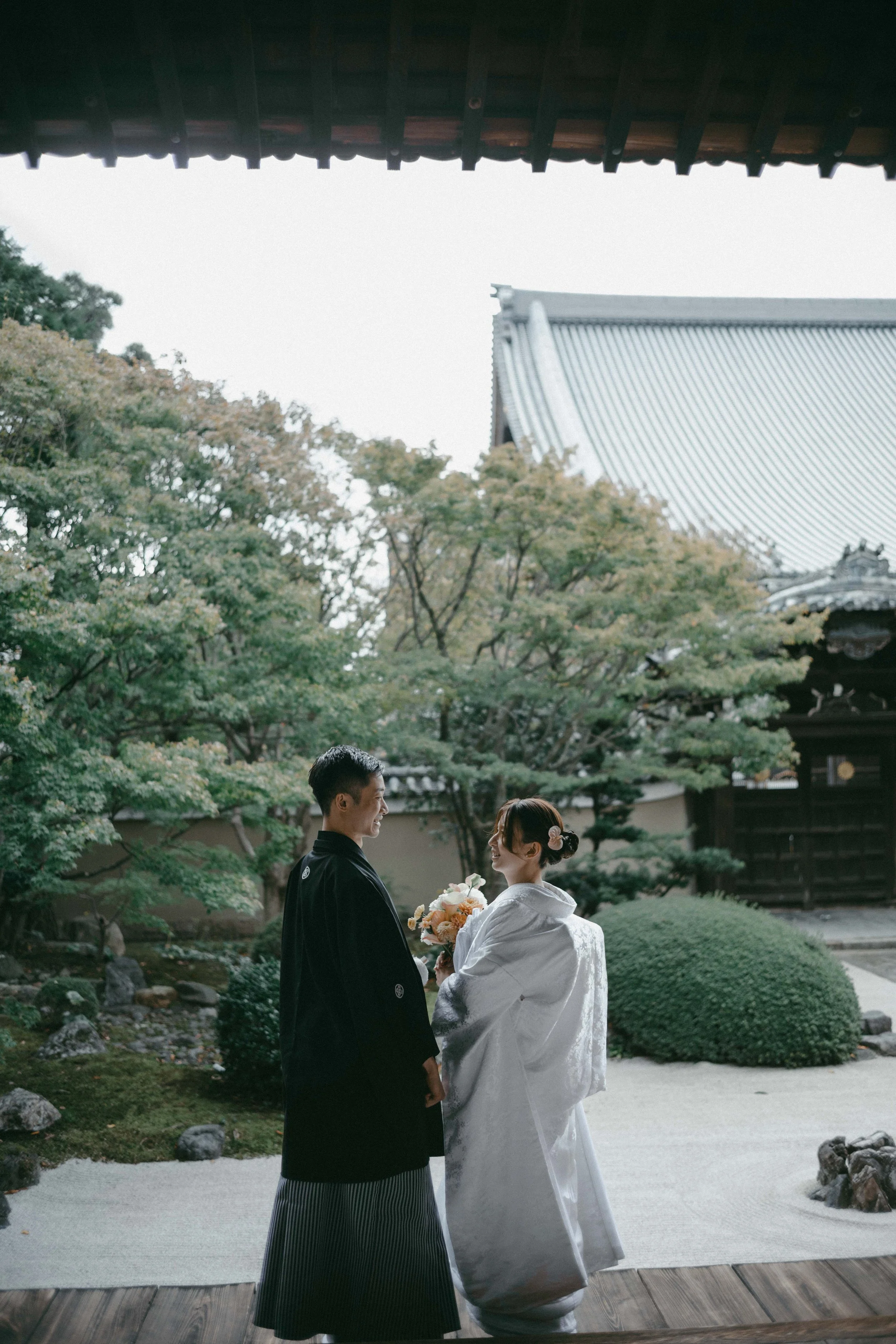 A couple dressed in traditional Japanese wedding attire standing outdoors in a Japanese garden, sharing a moment together. The groom wears a black kimono, and the bride wears a white kimono with a floral hair accessory. The background features lush g