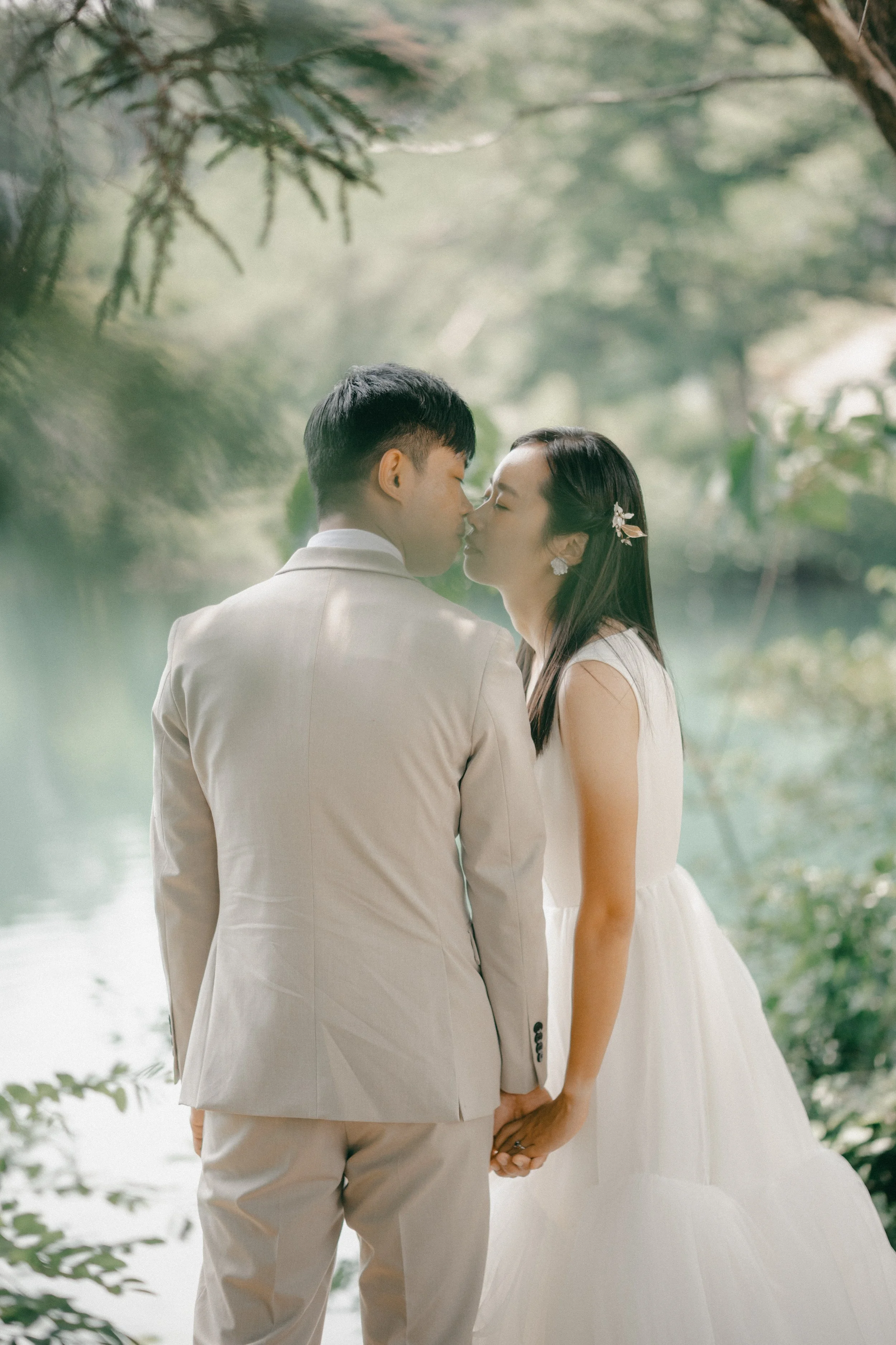 A couple dressed in wedding attire sharing a kiss and holding hands outdoors near a body of water, surrounded by greenery.