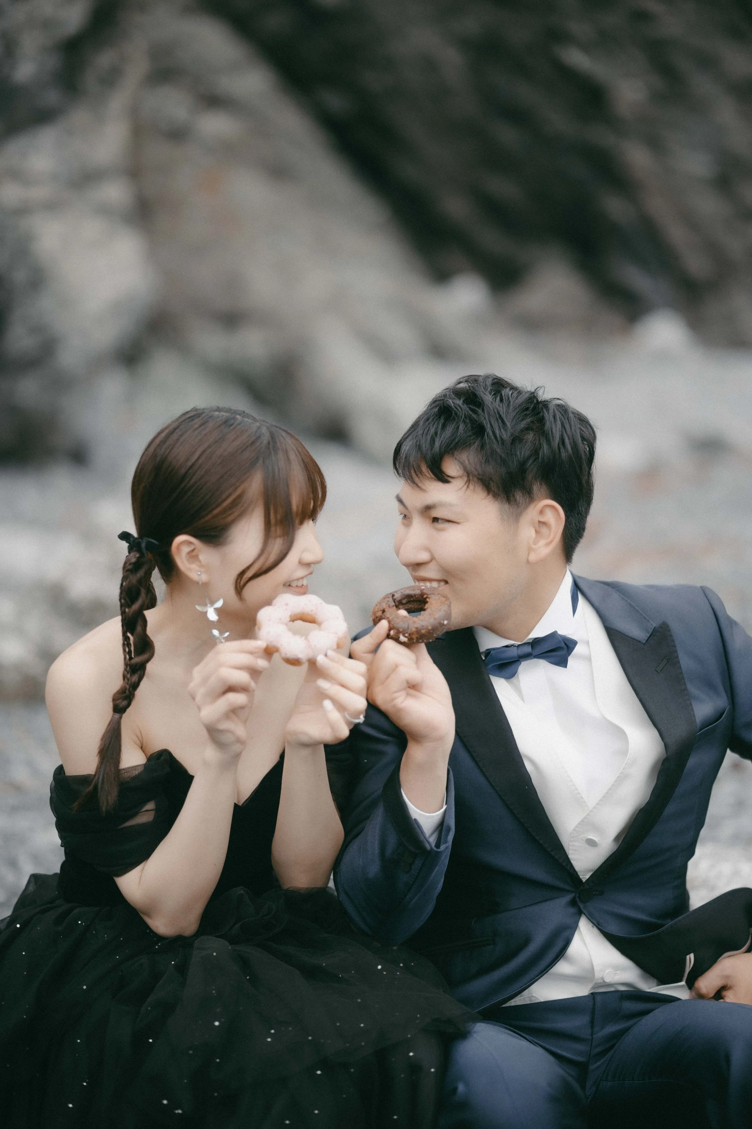 A couple dressed in formal attire, sitting on rocks near a beach, sharing a moment while holding donuts.