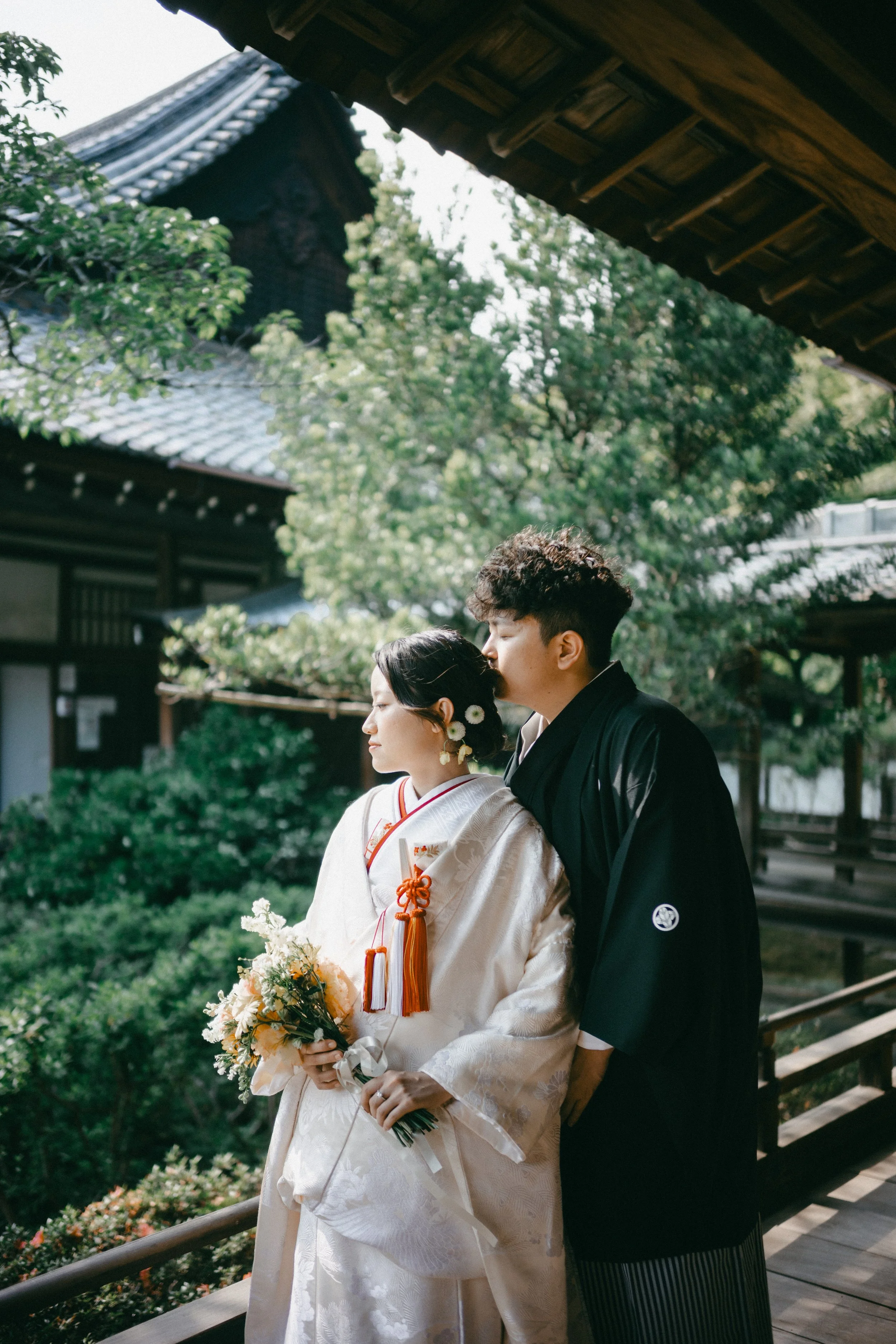 A Japanese bride and groom in traditional clothing standing outdoors on a porch of a traditional wooden building surrounded by lush greenery, with the groom kissing the bride's temple while she holds a bouquet of flowers.
