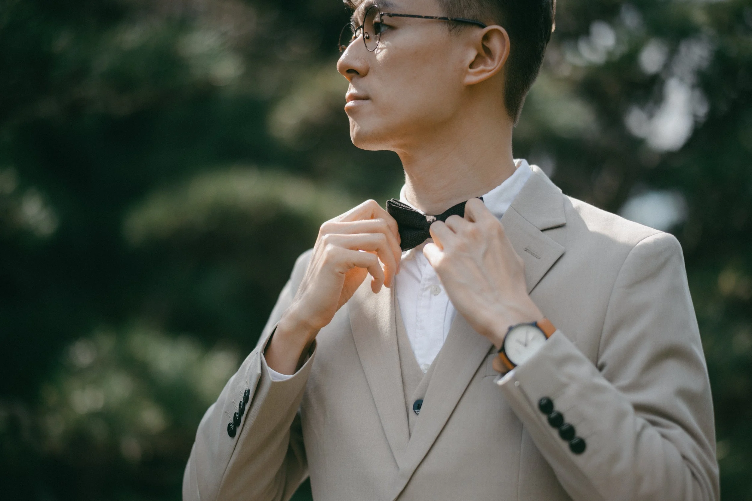 A man is adjusting a black bow tie outdoors. He is wearing glasses, a light beige suit jacket, a white shirt, and a wristwatch with a white face and brown leather strap.