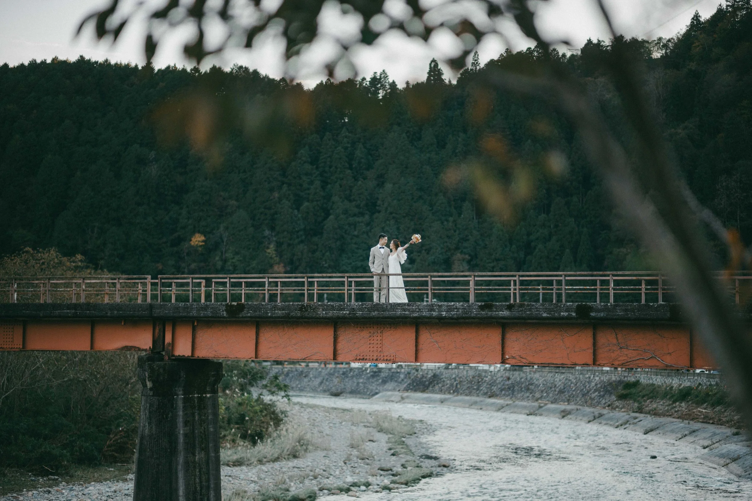 A bride and groom standing on a bridge with a forested mountain background, framed by blurred trees in the foreground.