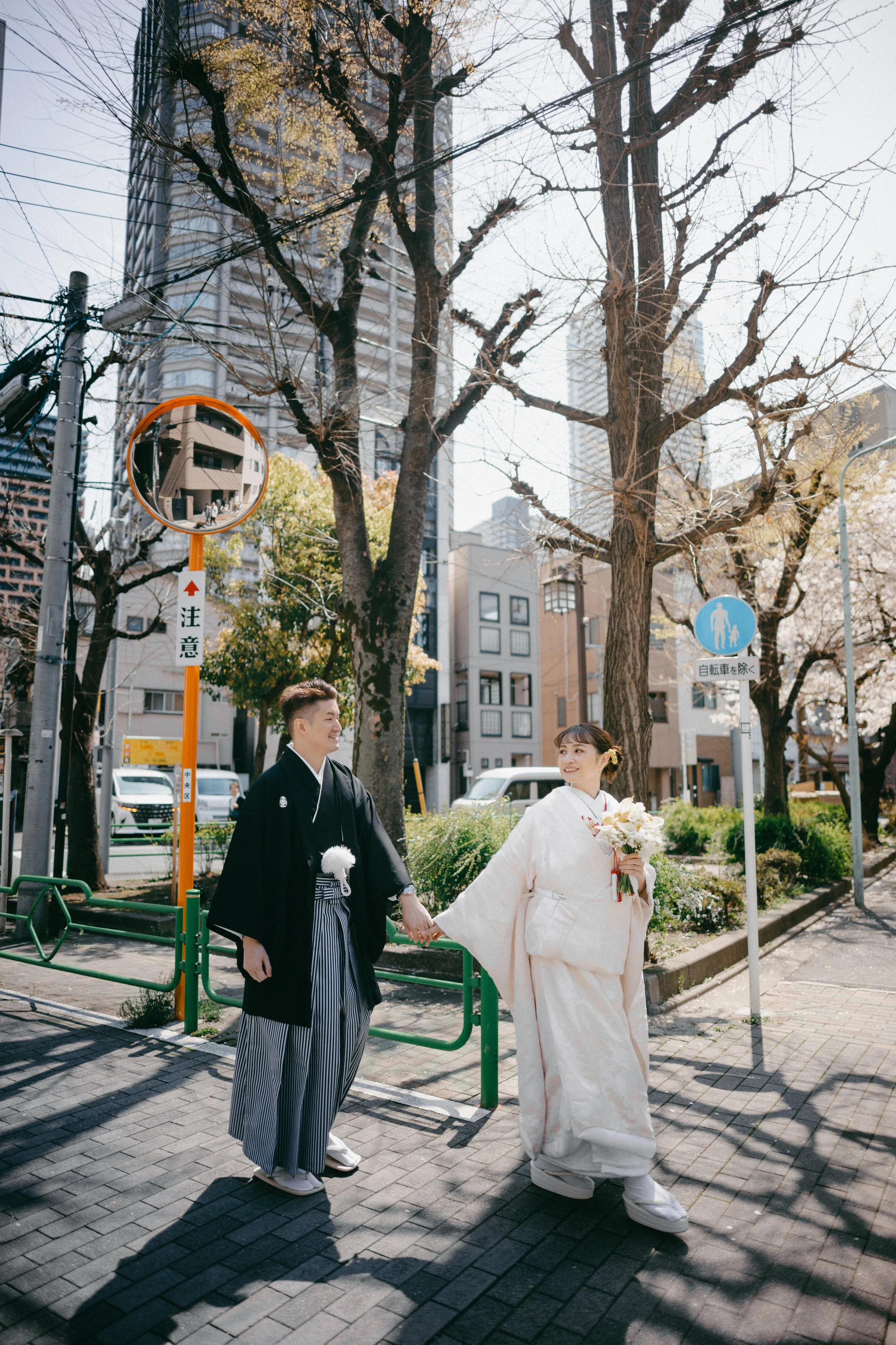 A bride and groom holding hands and walking outdoors on a sunny day in an urban area. The bride is wearing a traditional white wedding kimono and holding a bouquet, while the groom is dressed in a black kimono with striped hakama. There are leafless 