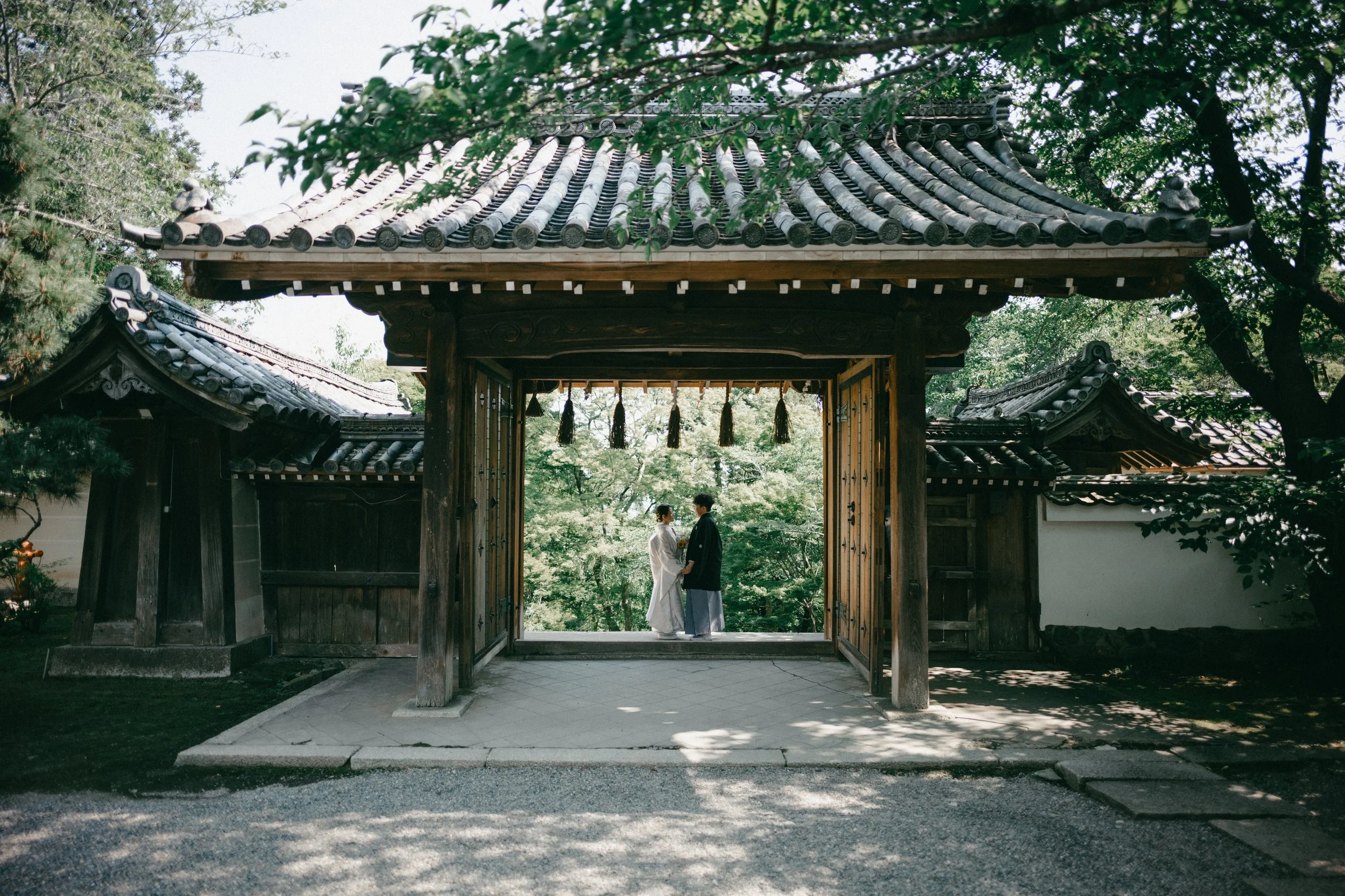 A traditional Japanese gate with a couple dressed in wedding attire standing under it, surrounded by lush green trees.