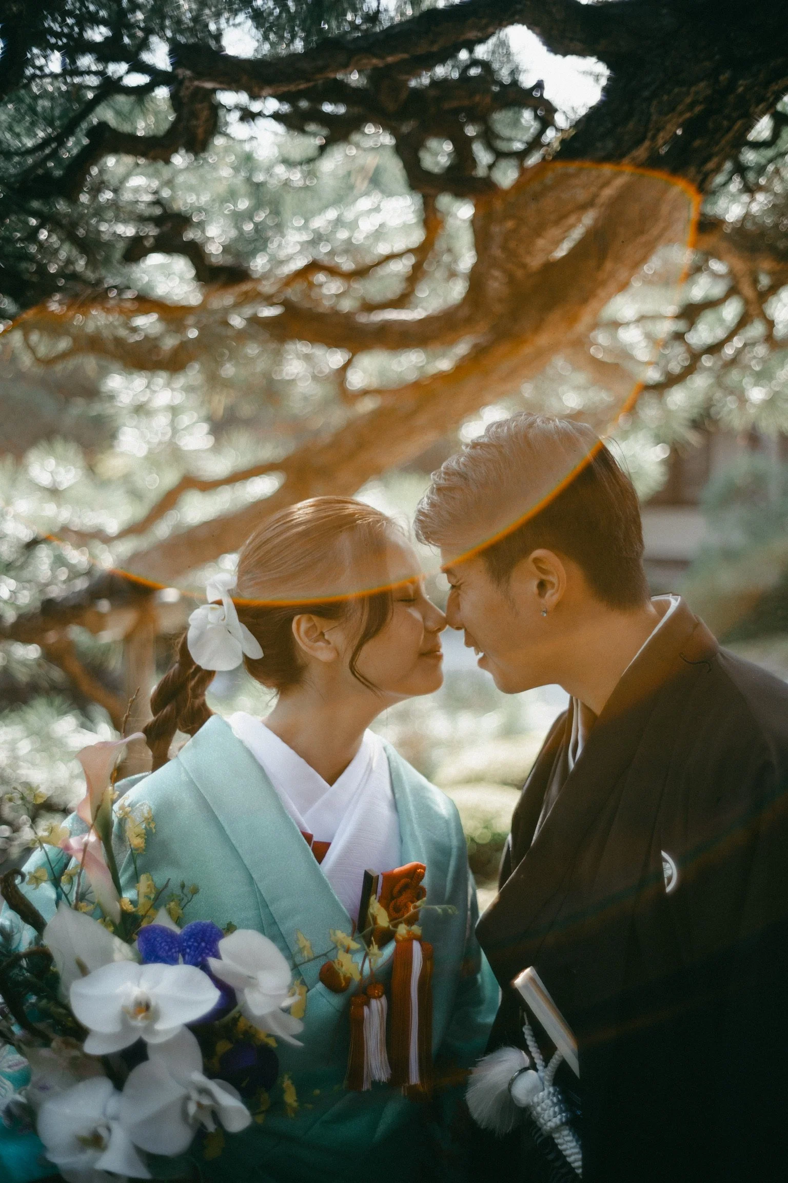 A couple in traditional Japanese clothing with foreheads touching, standing under a tree, holding flowers.