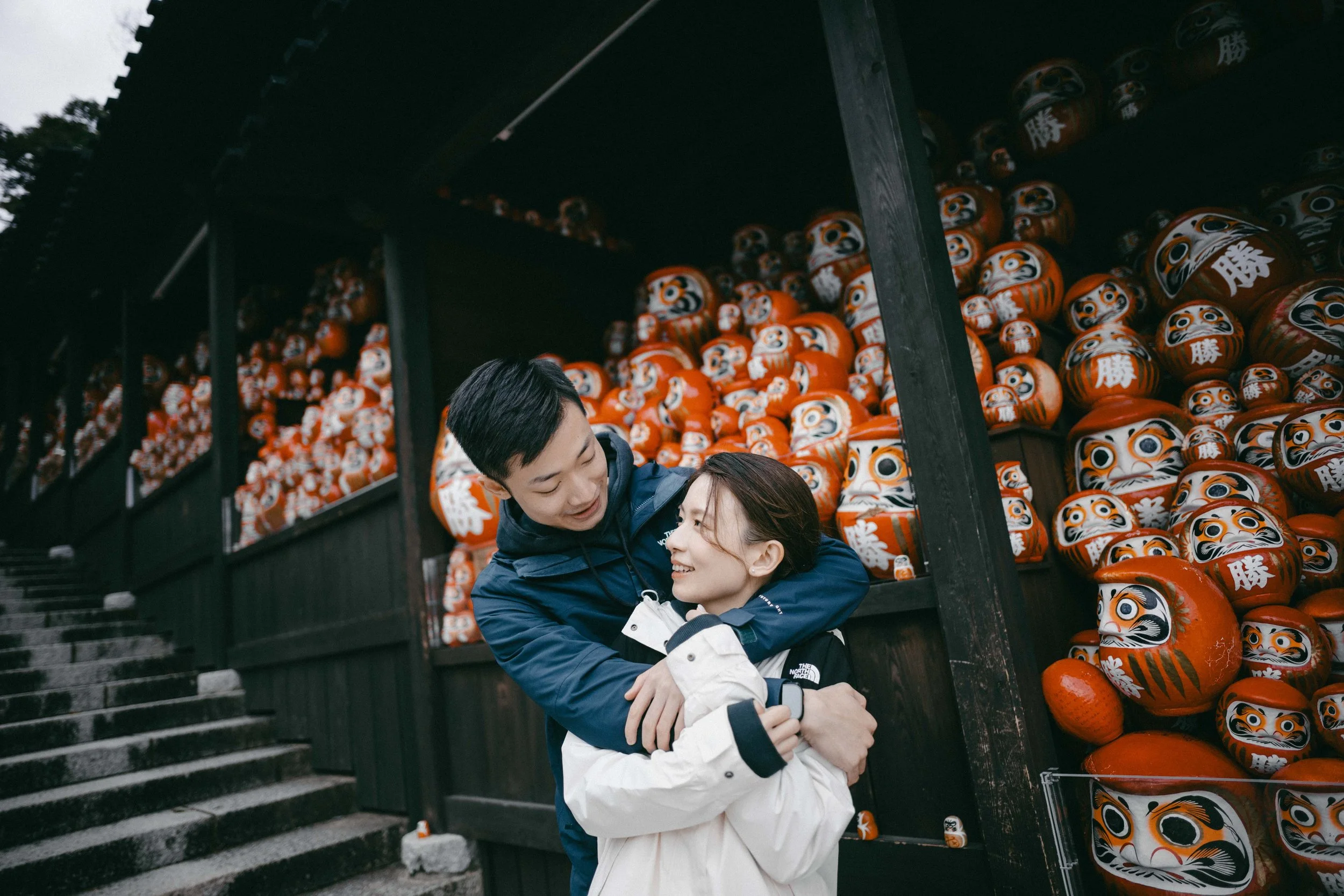 A smiling couple embracing each other outdoors with a display of orange daruma dolls in the background.